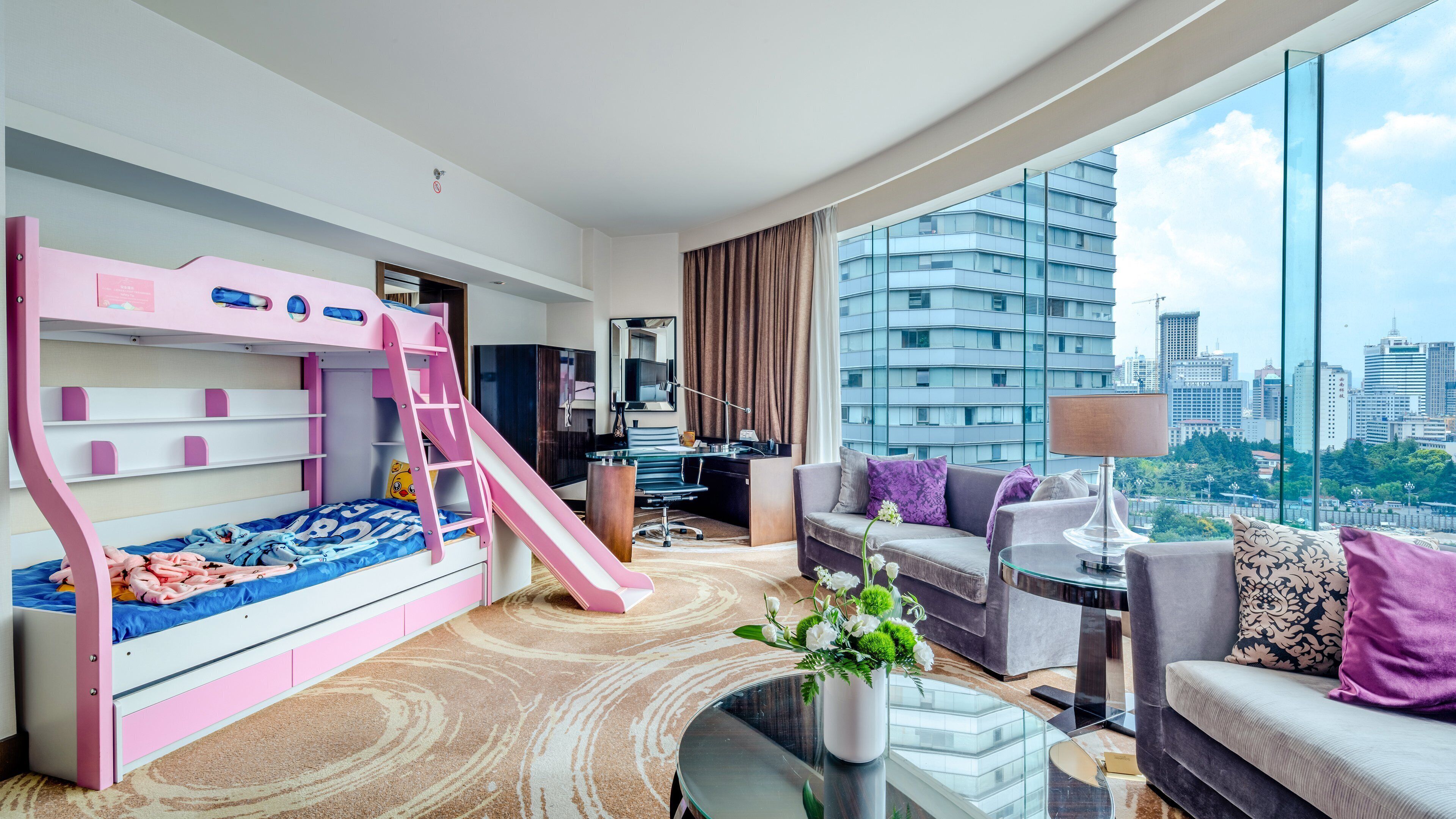 Habitación de hotel moderna con literas, escritorio, sofá y un amplio ventanal con vista a la ciudad.