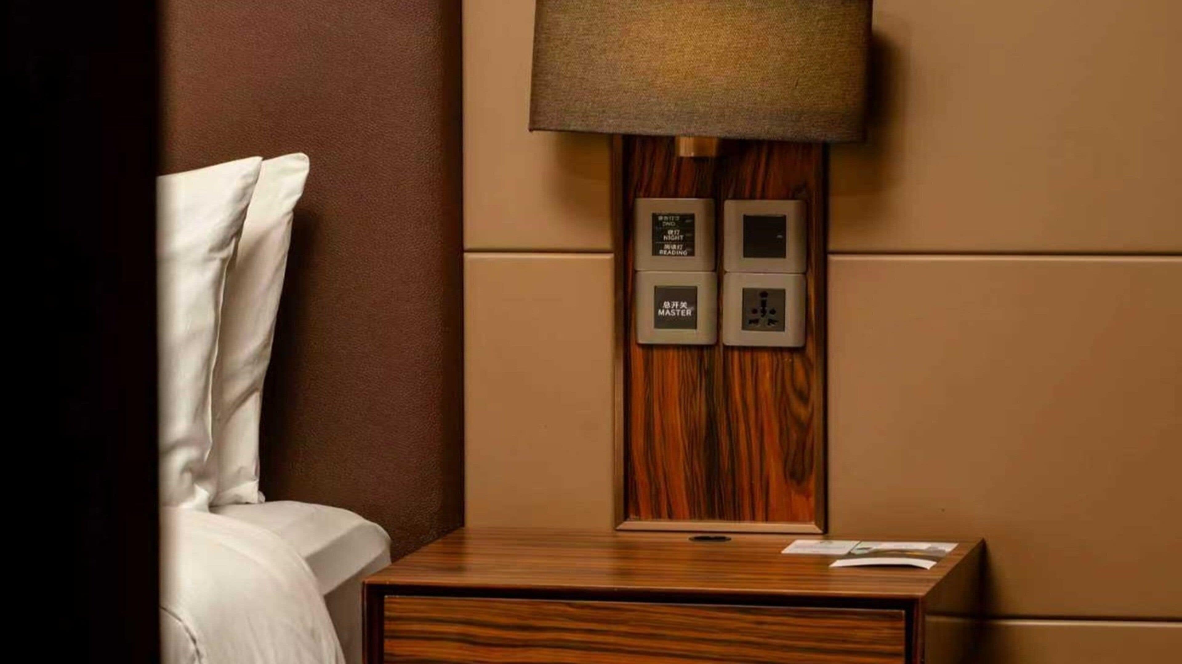 A hotel room with a wooden nightstand, a bed with white linens, and a wall-mounted lamp. There are two electrical outlets and a power strip on the wall.