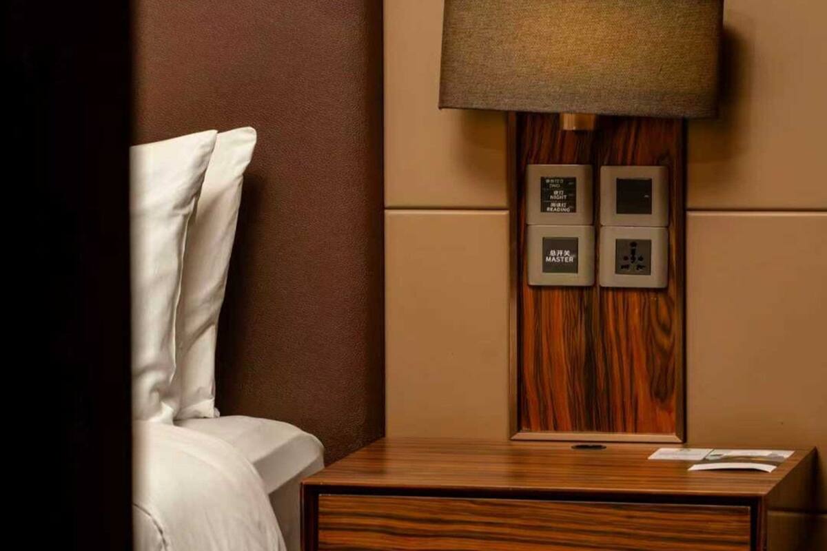 Una habitación de hotel con una mesita de noche de madera, una cama con ropa blanca y una lámpara de pared. Se observan dos tomas de corriente y una regleta en la pared.