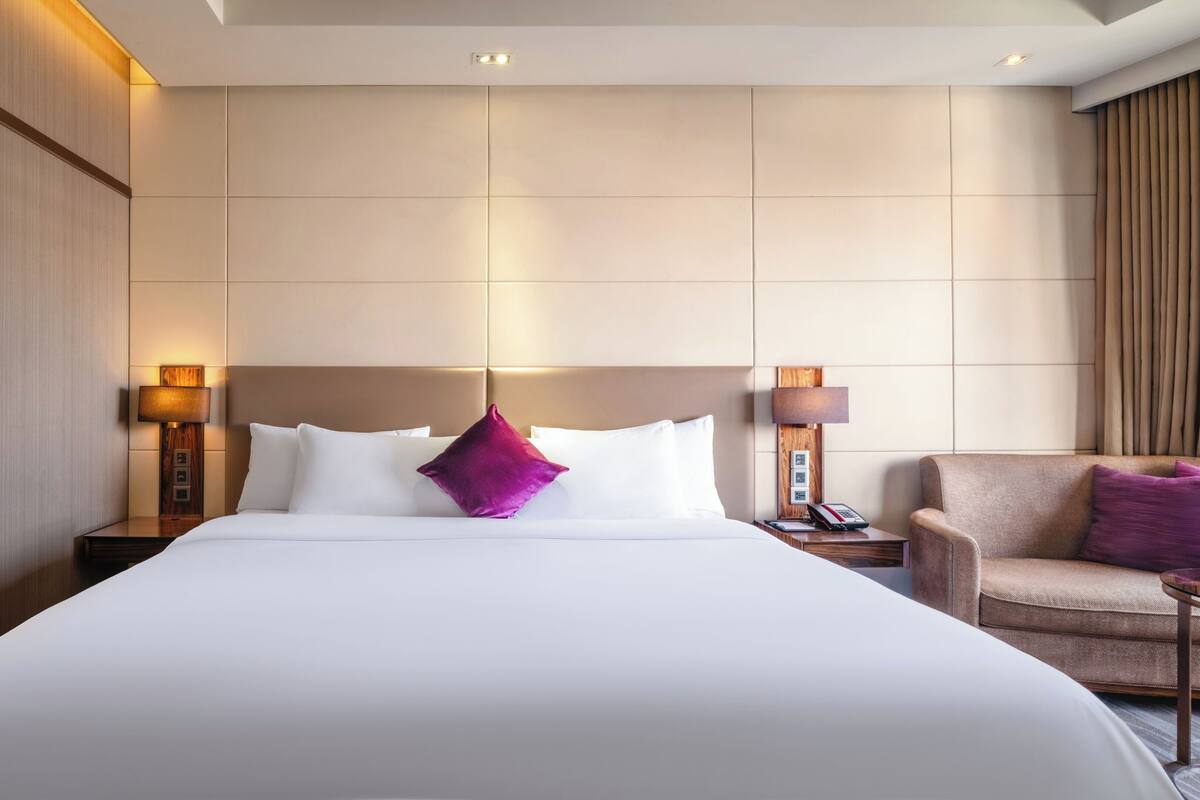 Habitación de hotel con una cama grande, dos mesitas de noche con lámparas, una silla y cabecera con iluminación integrada.