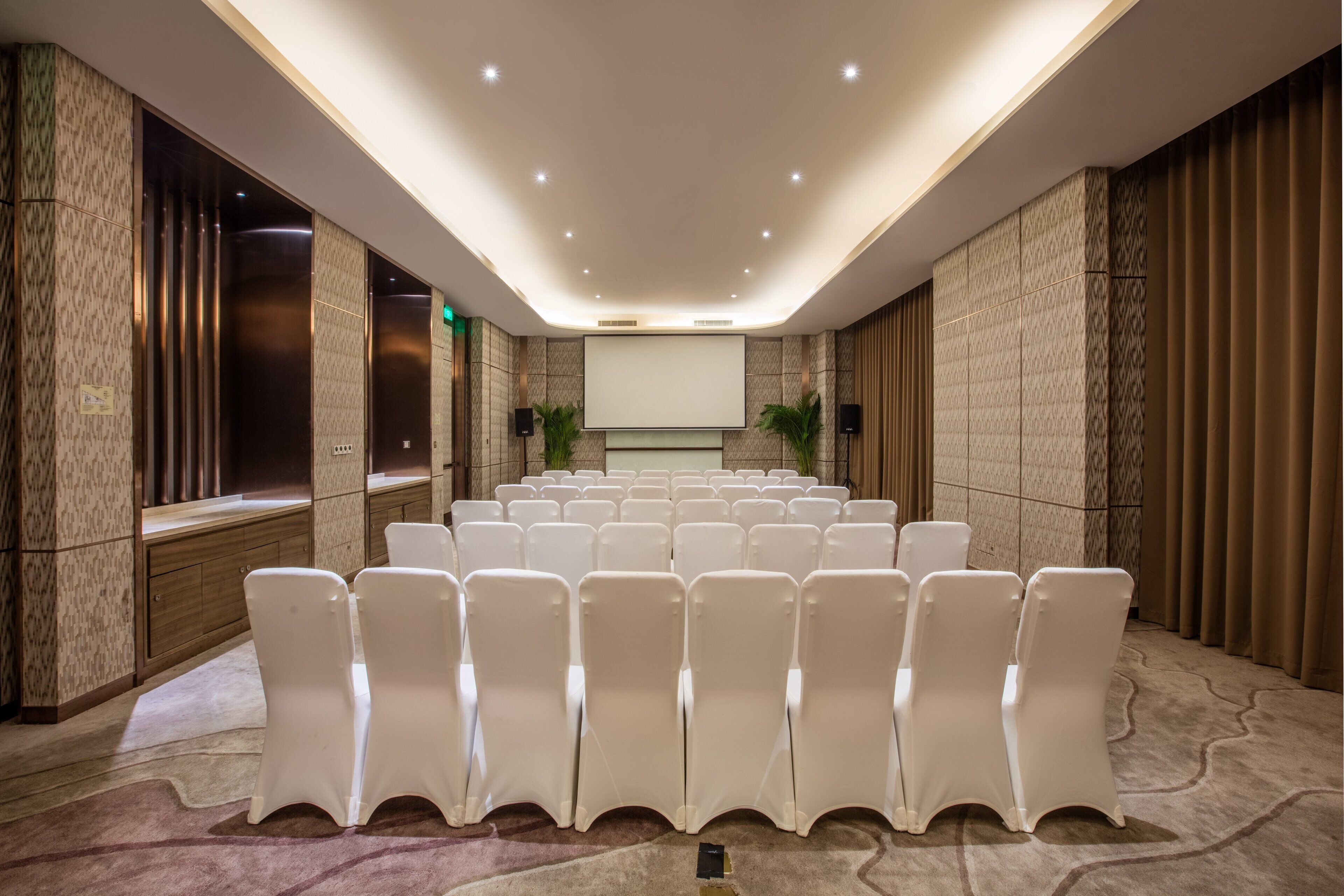 A conference room with rows of white chairs, a large screen, and a marble wall.