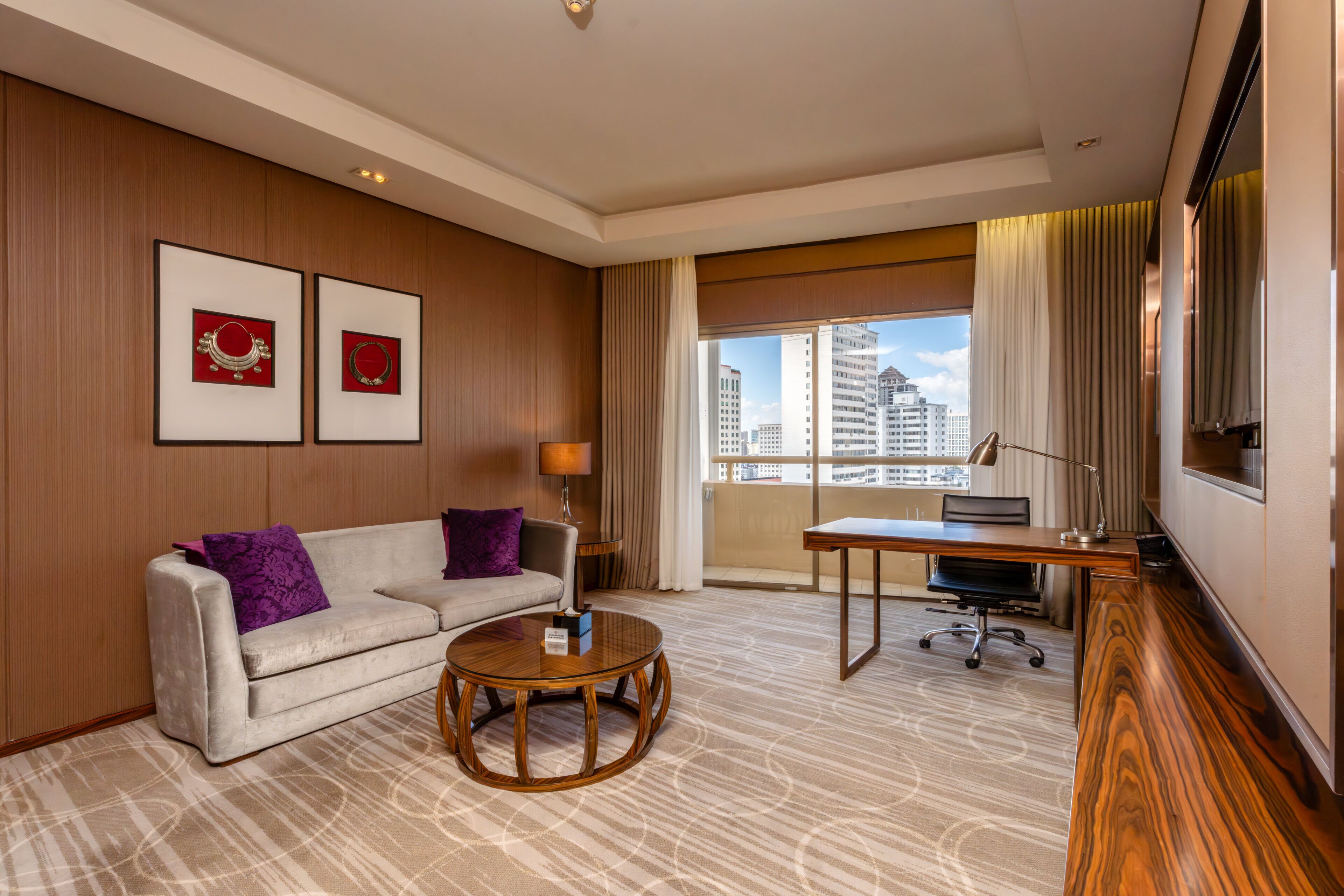 A hotel room with a sofa, a round coffee table, a desk, and a large window with a city view.