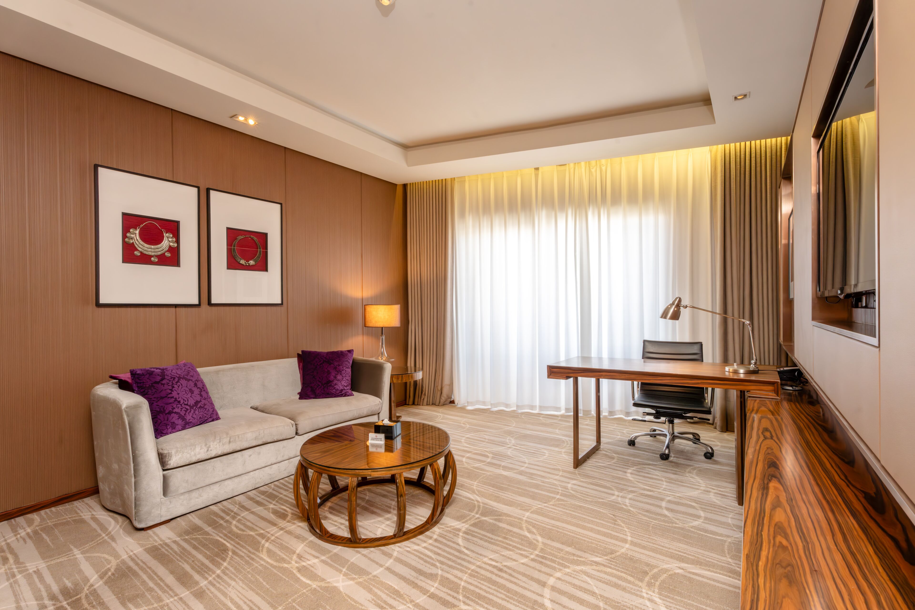 Une chambre d’hôtel comprenant un canapé, une table basse ronde, un bureau avec une chaise, un téléviseur et des œuvres d’art encadrées accrochées au mur.