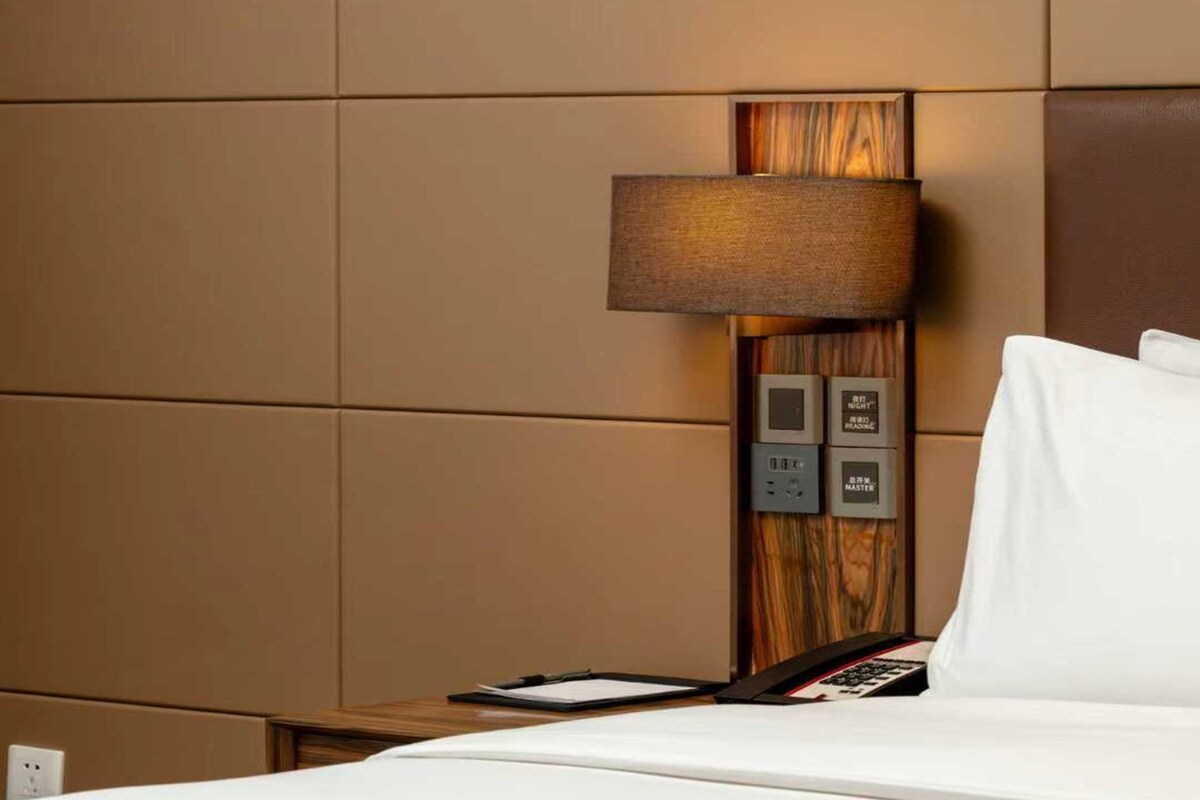 Una habitación de hotel con cama, mesita de noche, lámpara y panel de control con botones.