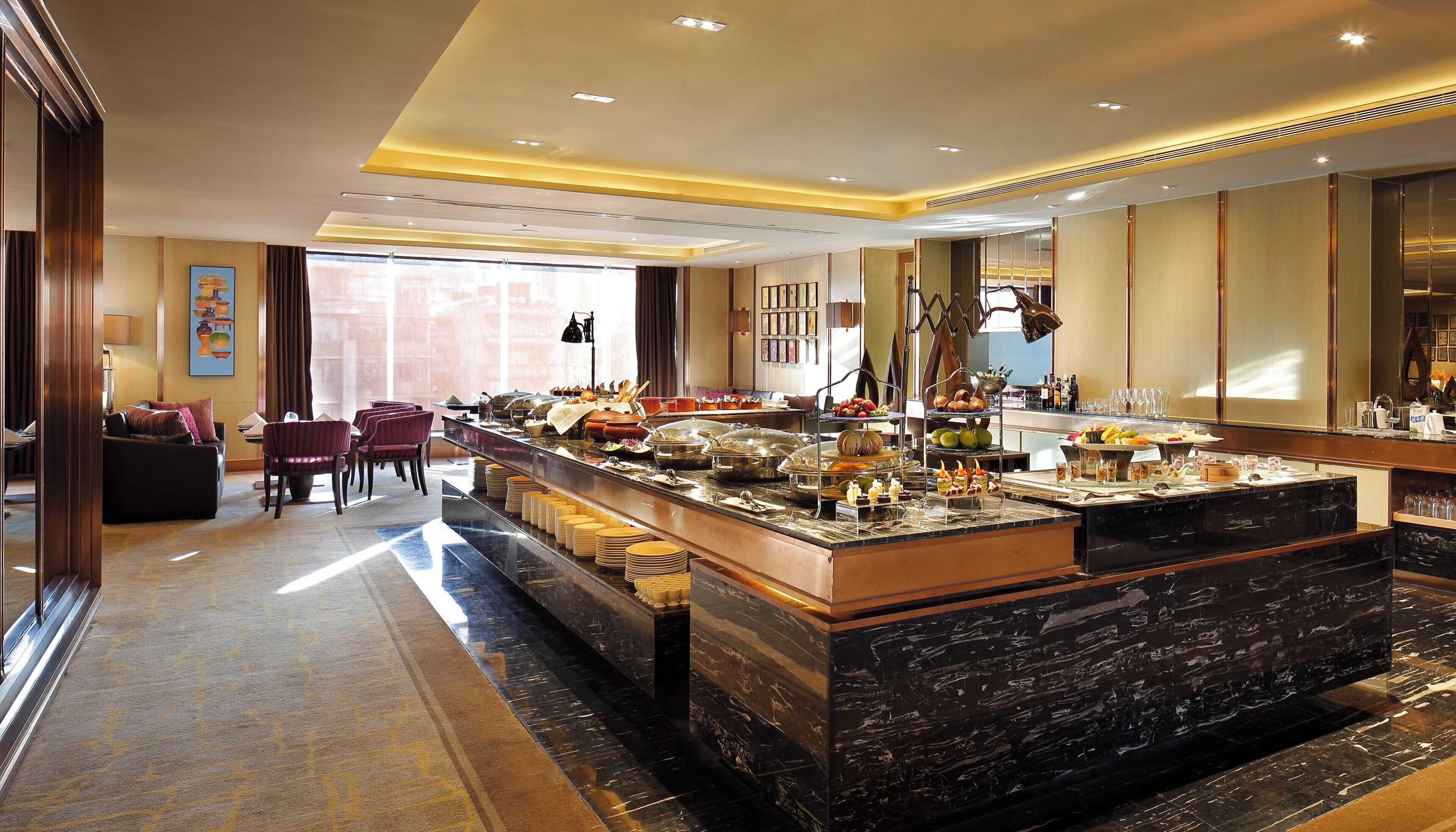 A hotel room with a large breakfast bar, a seating area with chairs, and a view of the city through large windows.
