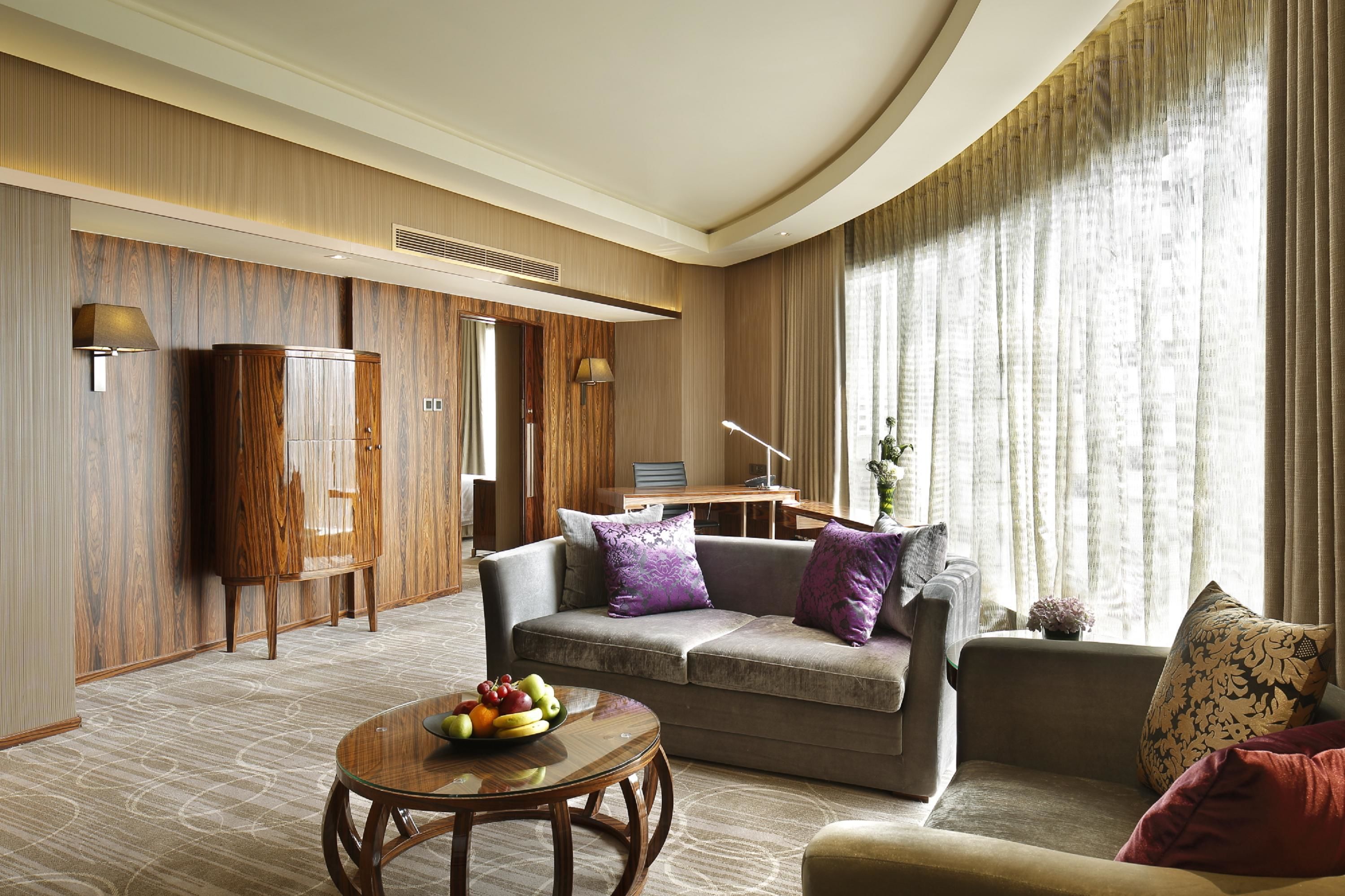 Una habitación de hotel moderna con sofá, una mesa redonda con fruta, un armario de madera y un ventanal con cortinas.