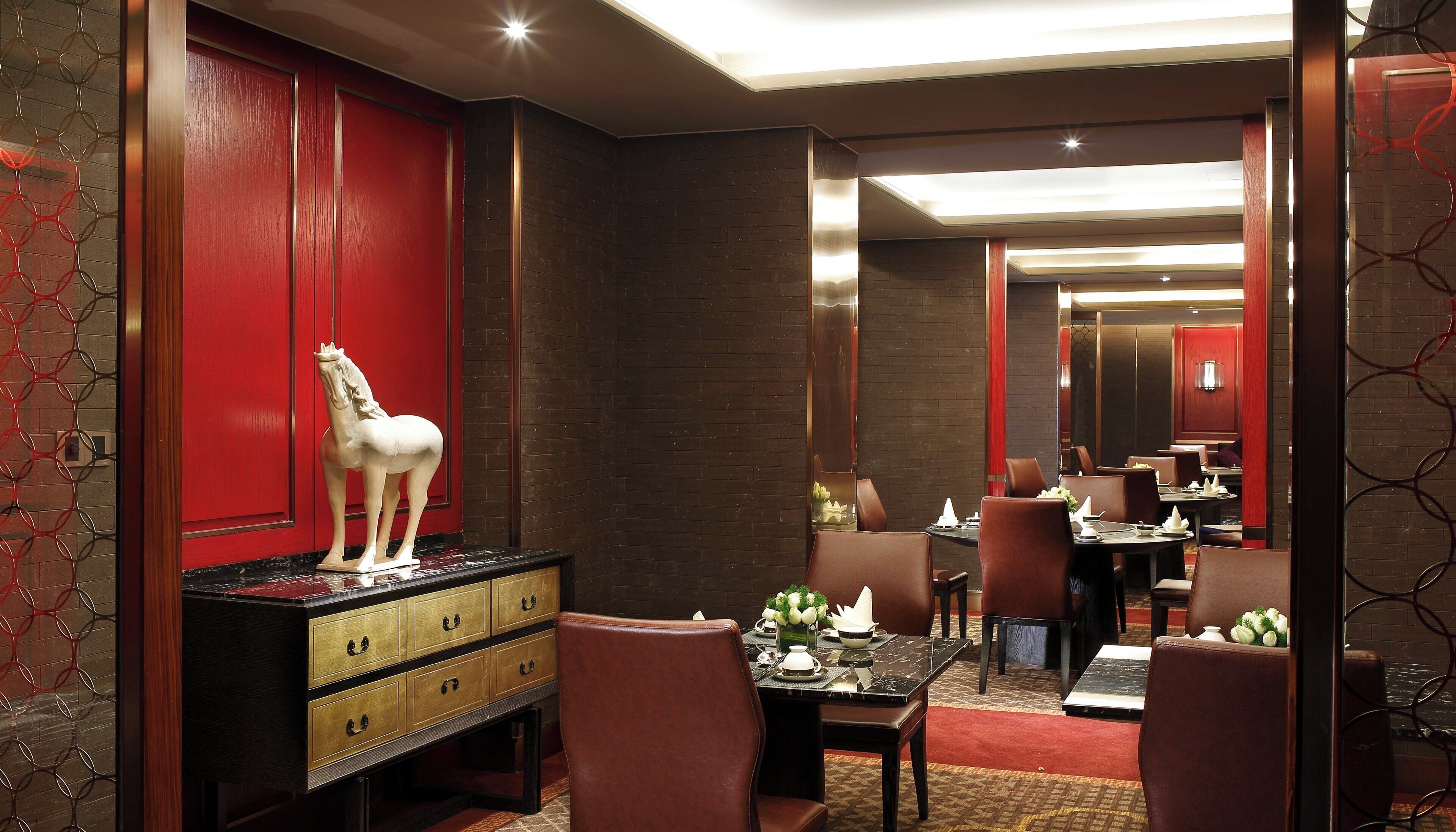 Un hall d’hôtel avec un mur rouge, un meuble en bois doté de tiroirs et une statue de cheval. On y trouve plusieurs tables et chaises disposées pour accueillir les clients.