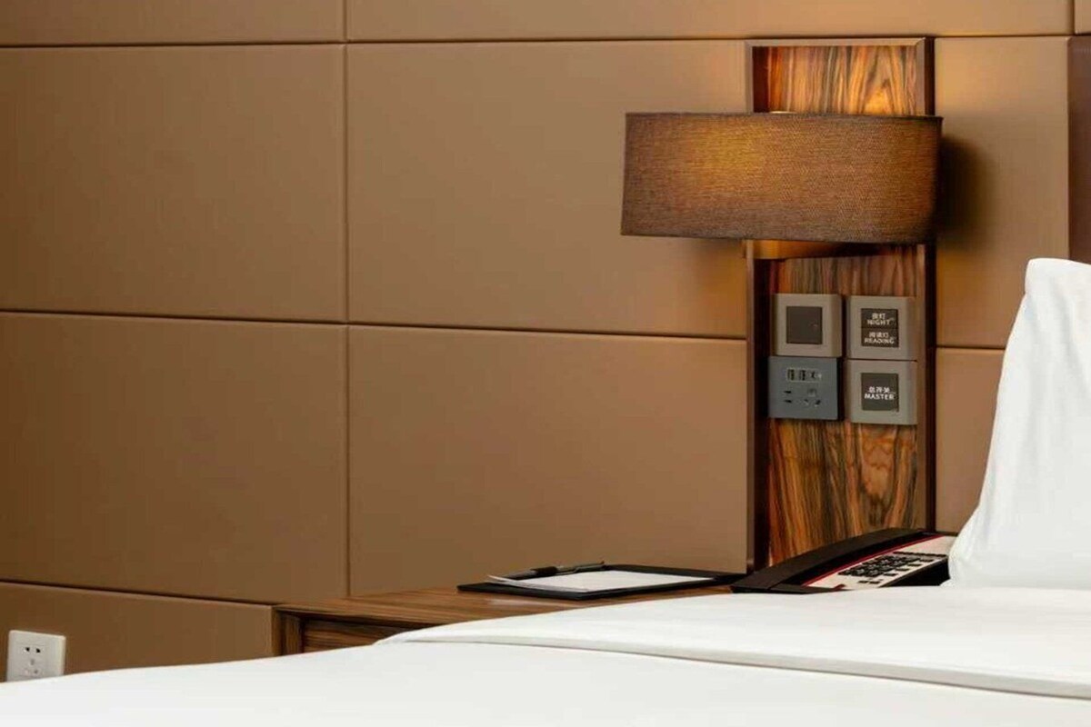 Habitación de hotel con cama, mesita de noche, lámpara y panel de control con botones.
