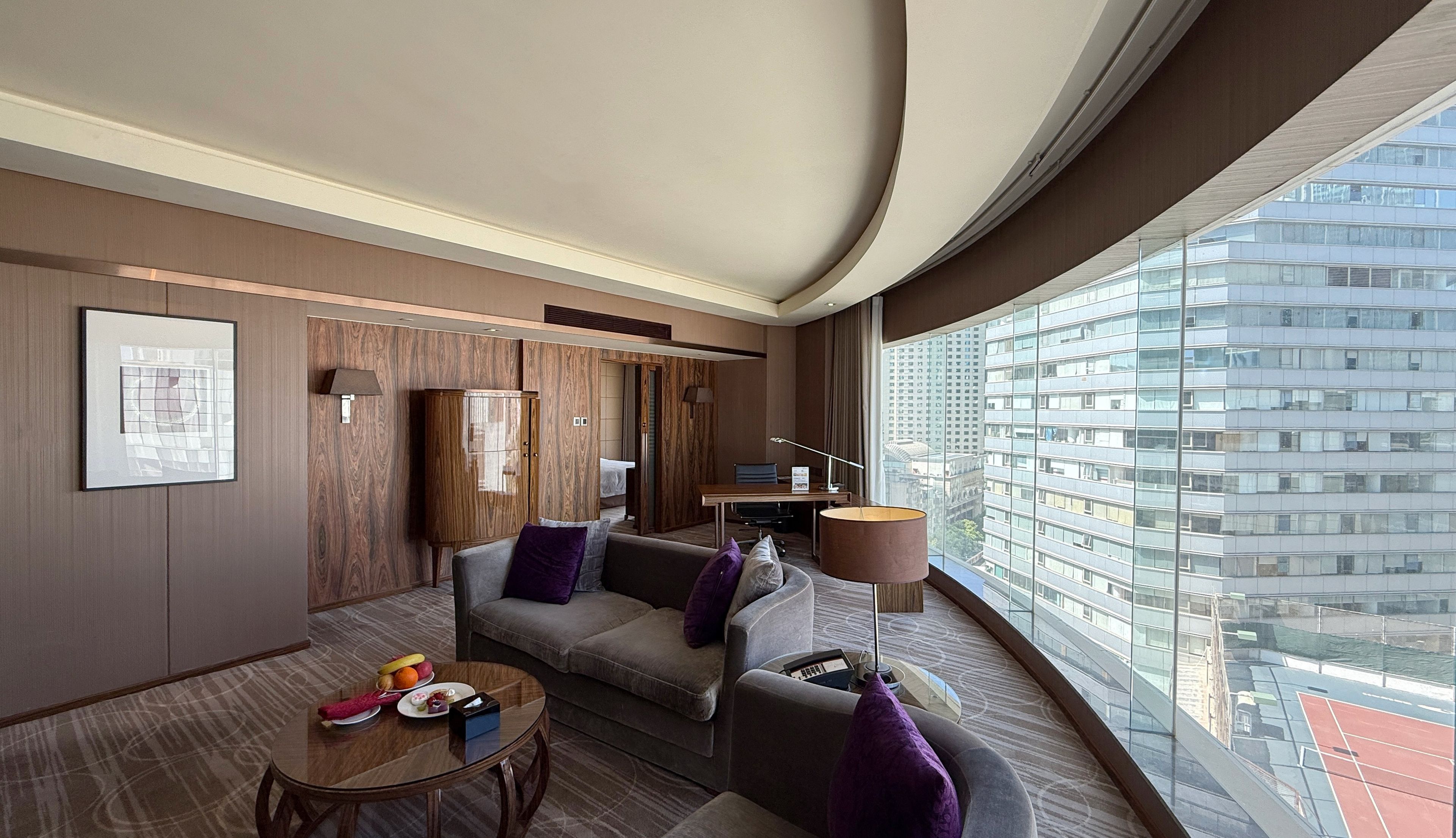 A modern hotel room with a curved ceiling, a large window with a city view, a sofa, a small table with fruit, and a cabinet.