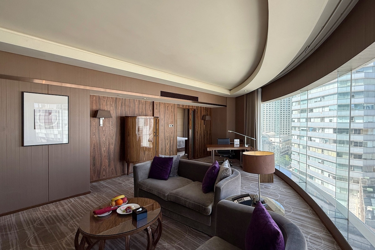 Een moderne hotelkamer met een gebogen plafond, een groot raam met uitzicht op de stad, een bank, een klein tafeltje met fruit en een kast.
