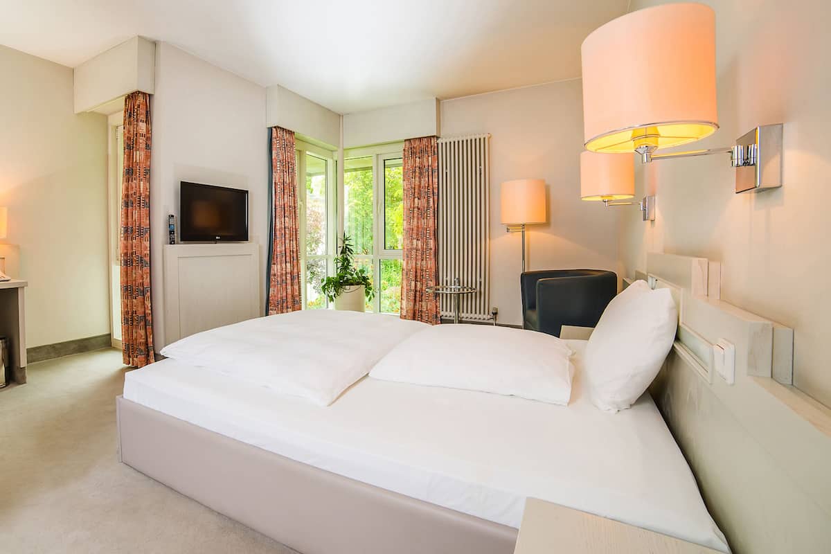 Ein Hotelzimmer mit einem großen Bett, einem Schreibtisch, einem Stuhl, einem Fernseher und einem Vorhangfenster.