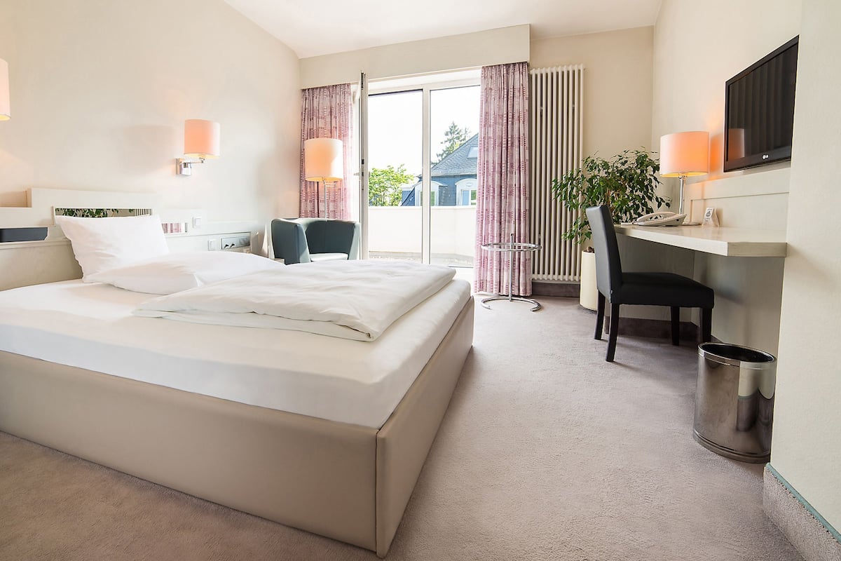 Ein Hotelzimmer mit einem großen Bett, einem Schreibtisch, einem Stuhl, einer Mülleimer und einem Fenster mit Vorhängen.