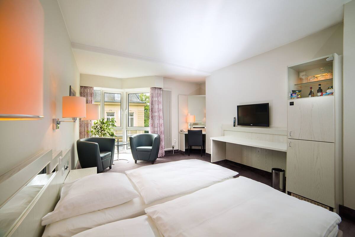 Ein Hotelzimmer mit Bett, Schreibtisch, Stuhl, Fernseher und einem Fenster mit Vorhängen.
