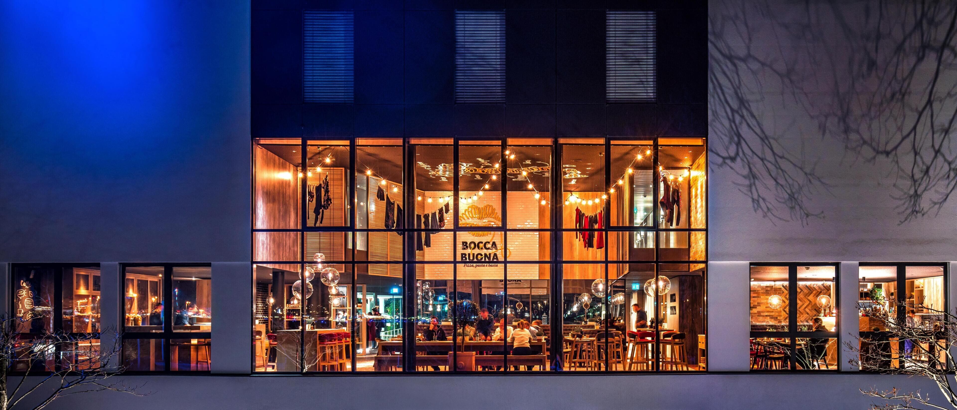 A restaurant with a glass facade, interior lighting, and visible seating areas.