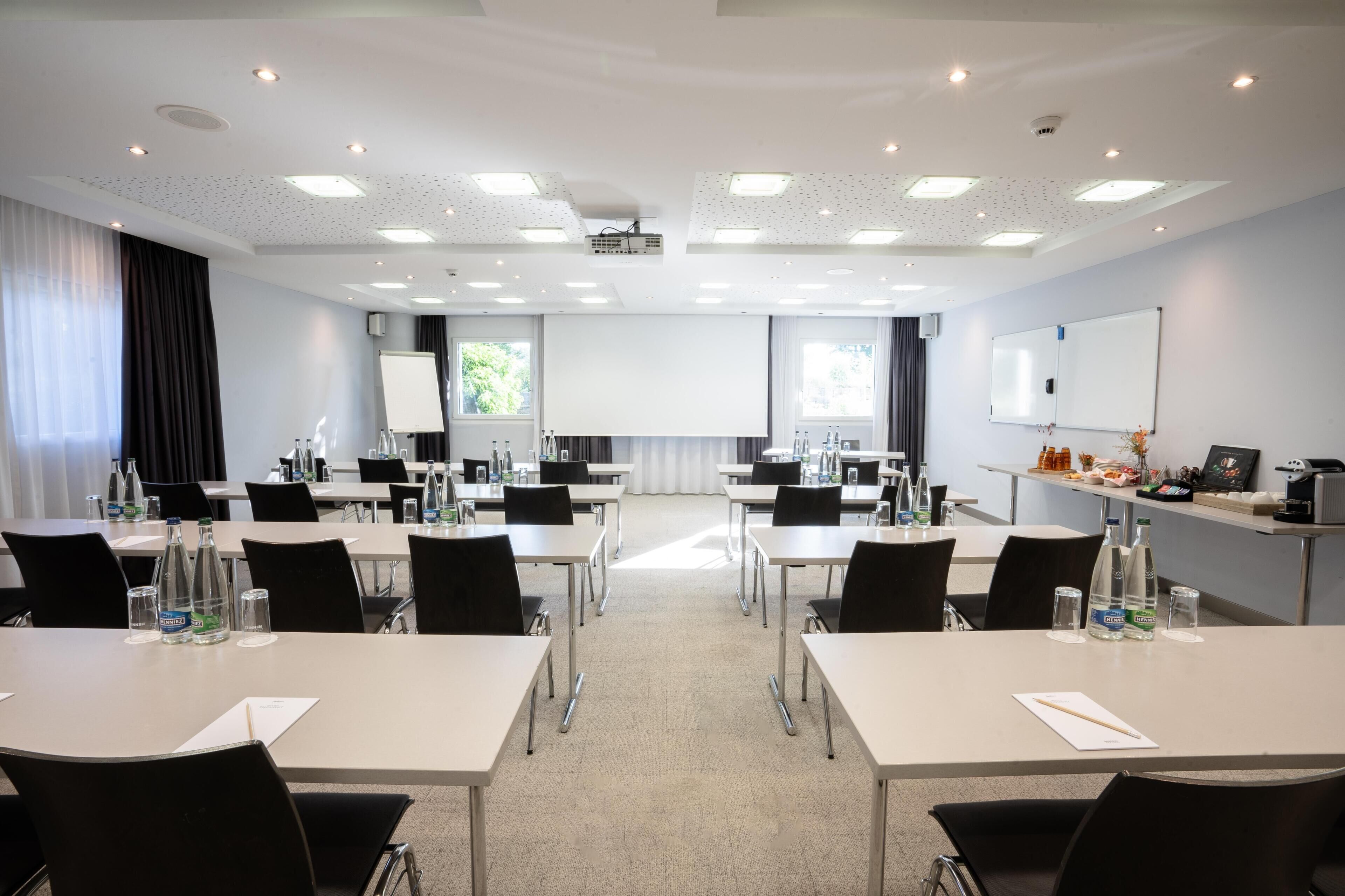 Ein Konferenzraum mit Tischen, Stühlen, einem Projektor, einer Tafel und einem Fenster.