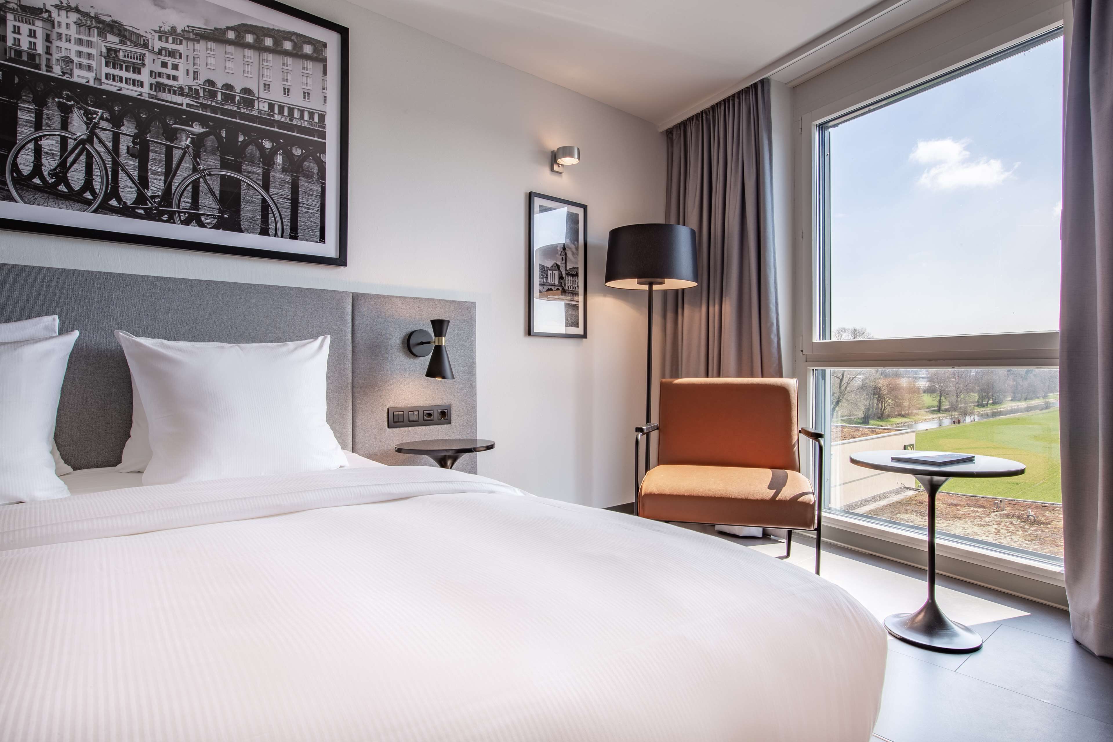 A hotel room with a large bed, a chair, a small table, a lamp, and a view of a park through a window.