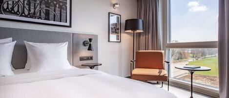 A hotel room with a large bed, a chair, a small table, a lamp, and a view of a park through a window.