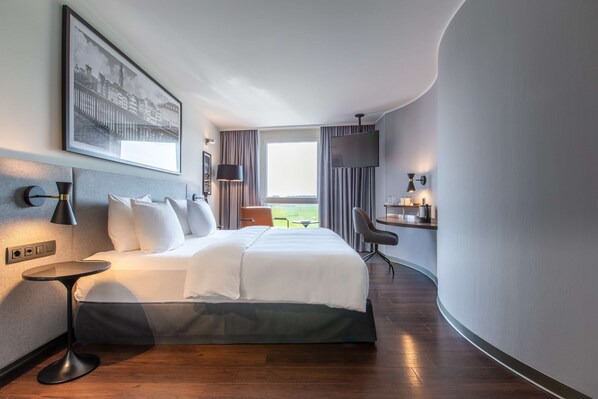 A modern hotel room with a large bed, a small table, a TV, and a view of the outdoors.