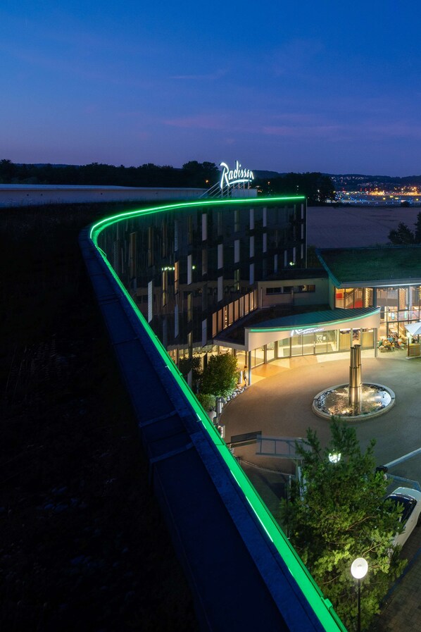 Un hotel con un letrero verde neón curvo, un patio central con una fuente y vistas de la ciudad al fondo.