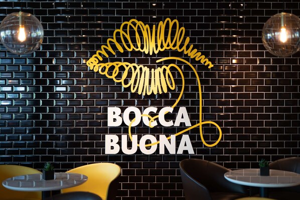 Un muro de ladrillo negro con un letrero de neón que dice "BOCCA BUONA" y dos mesas redondas con sillas.
