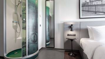 A modern bathroom with a glass-enclosed shower, a round mirror, and a bed with white linens.