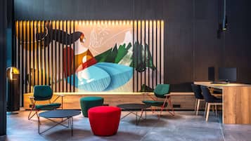A modern lounge area with a wooden desk, green and red chairs, and a colorful abstract mural on the wall.