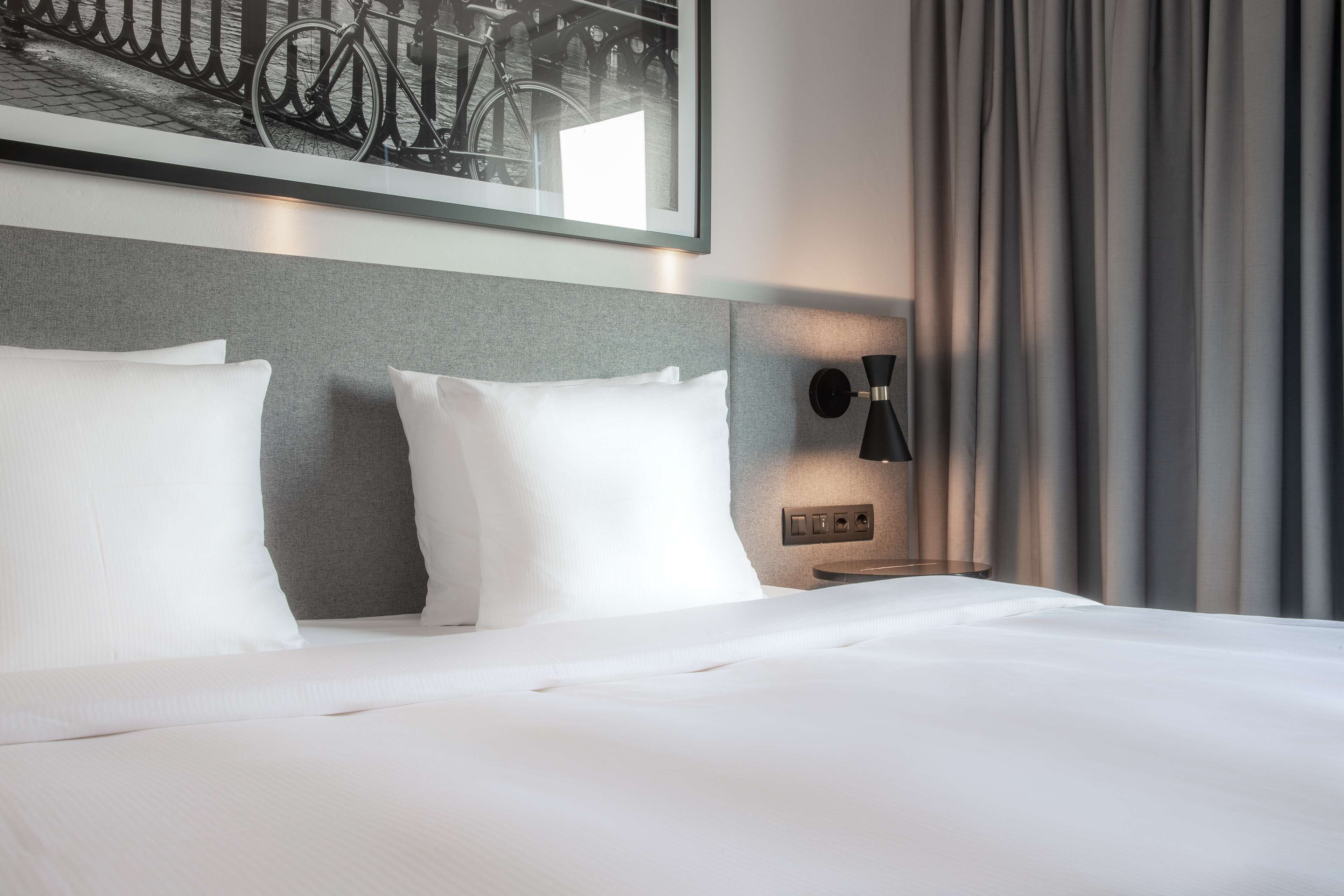 A neatly made bed with white linens, a grey headboard, and a wall-mounted lamp.