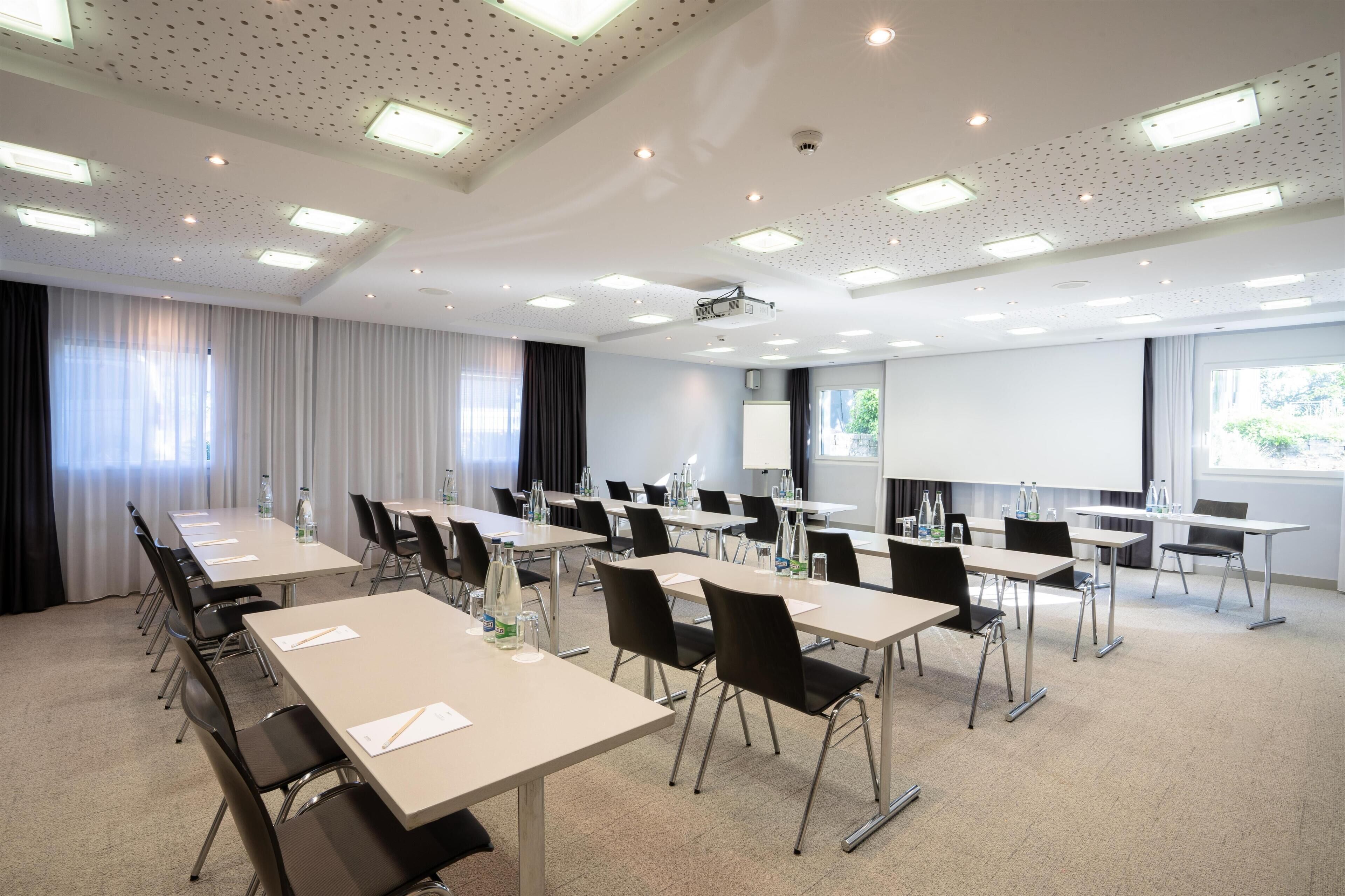 A conference room with multiple tables, chairs, a projector screen, and a large window.