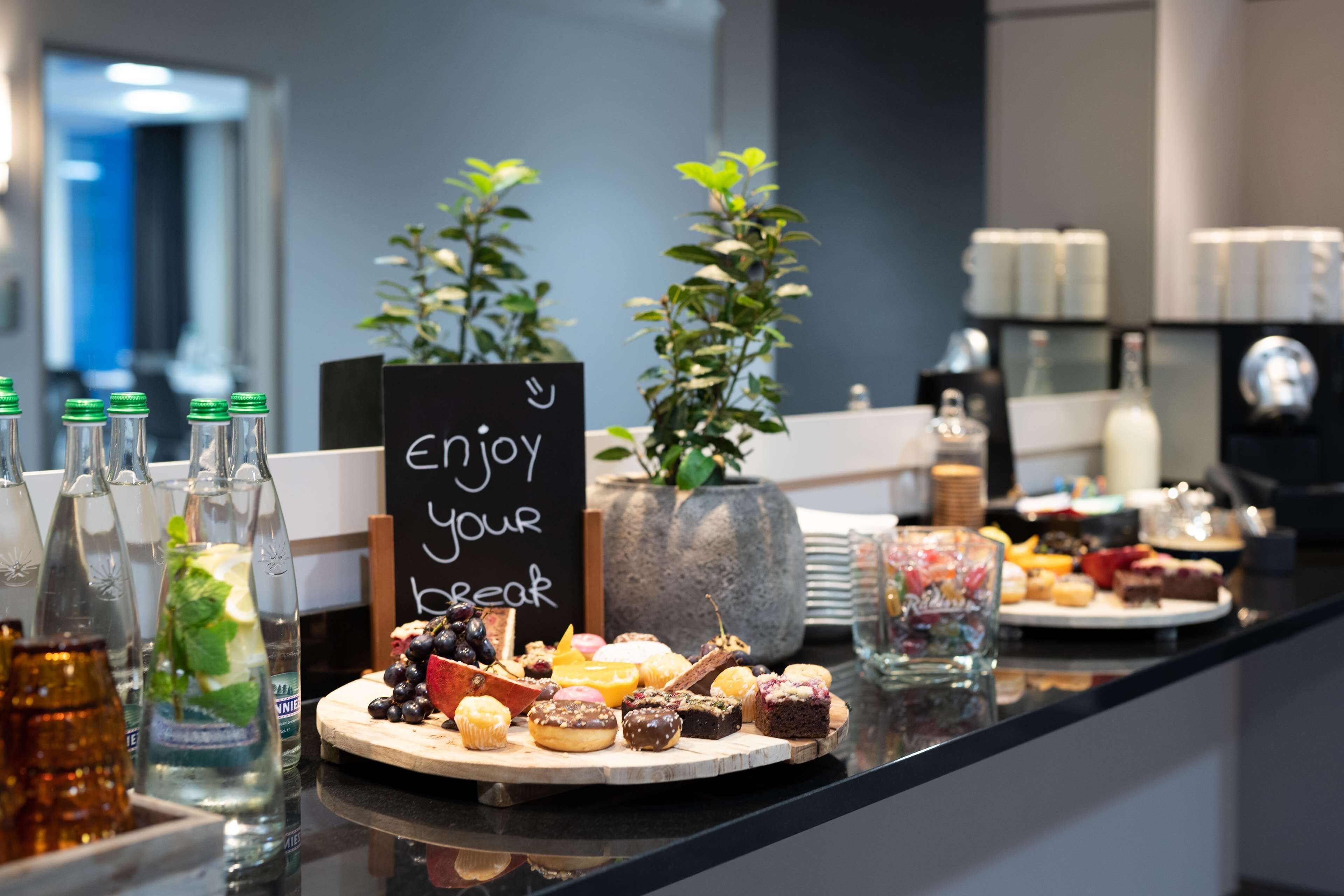 Ein Frühstücksbuffet mit verschiedenen Backwaren und Getränken, ein Schild mit der Aufschrift 'Genießen Sie Ihre Pause' sowie zwei Topfpflanzen.