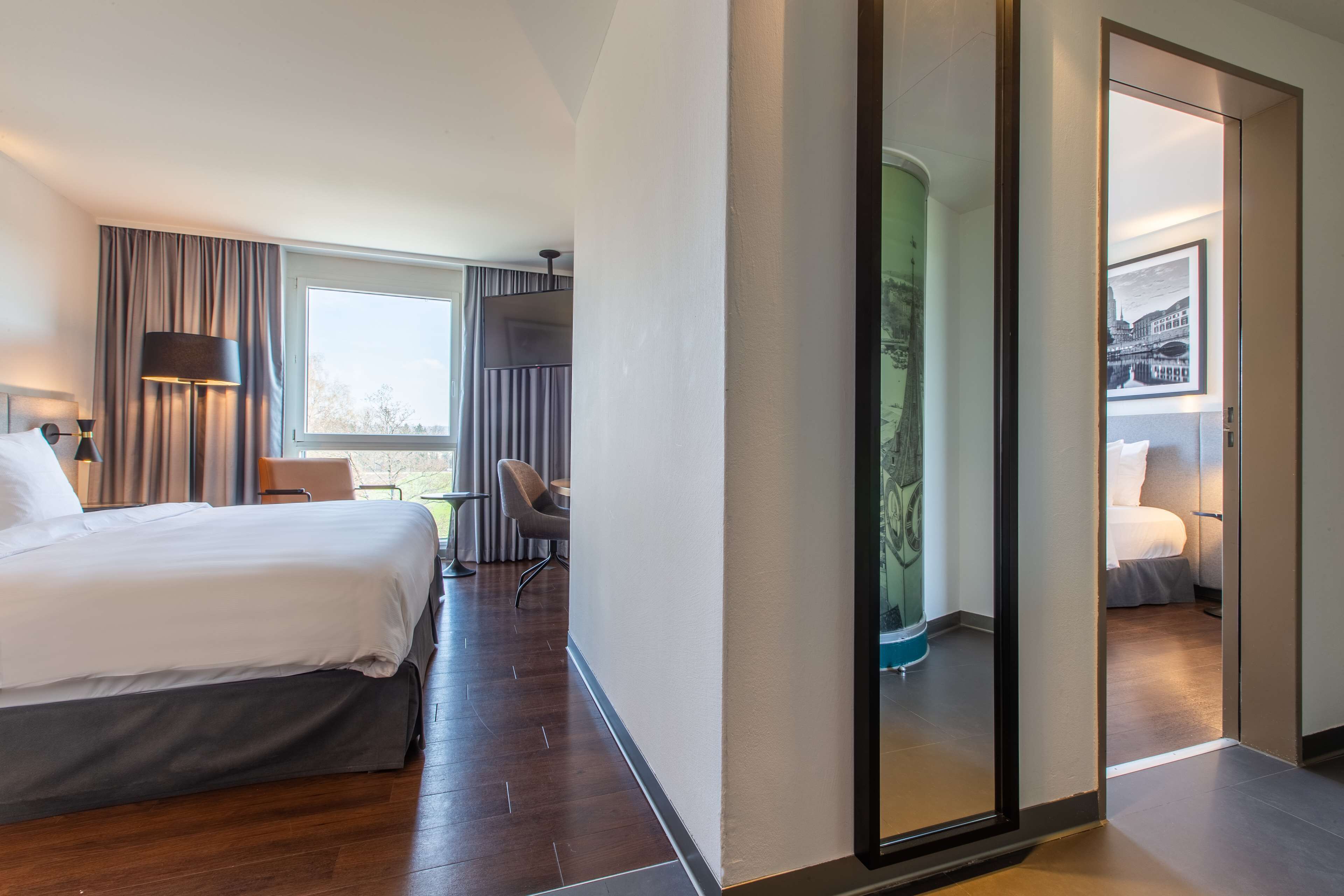 A hotel room with a large bed, a desk, a chair, a lamp, and a view of the outdoors.