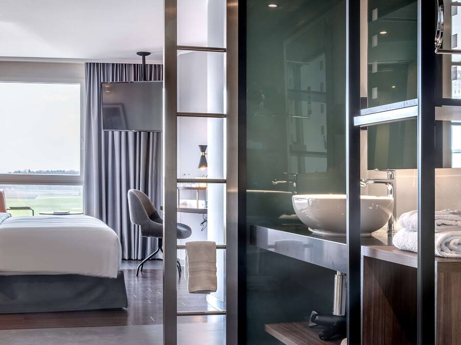 Habitación de hotel moderna con una cama grande, un escritorio, una silla, un baño con lavamanos y espejo, y vistas al exterior.
