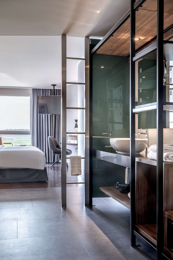 Habitación de hotel moderna con una cama grande, un escritorio, una silla, un baño con lavamanos y espejo, y vistas al exterior.