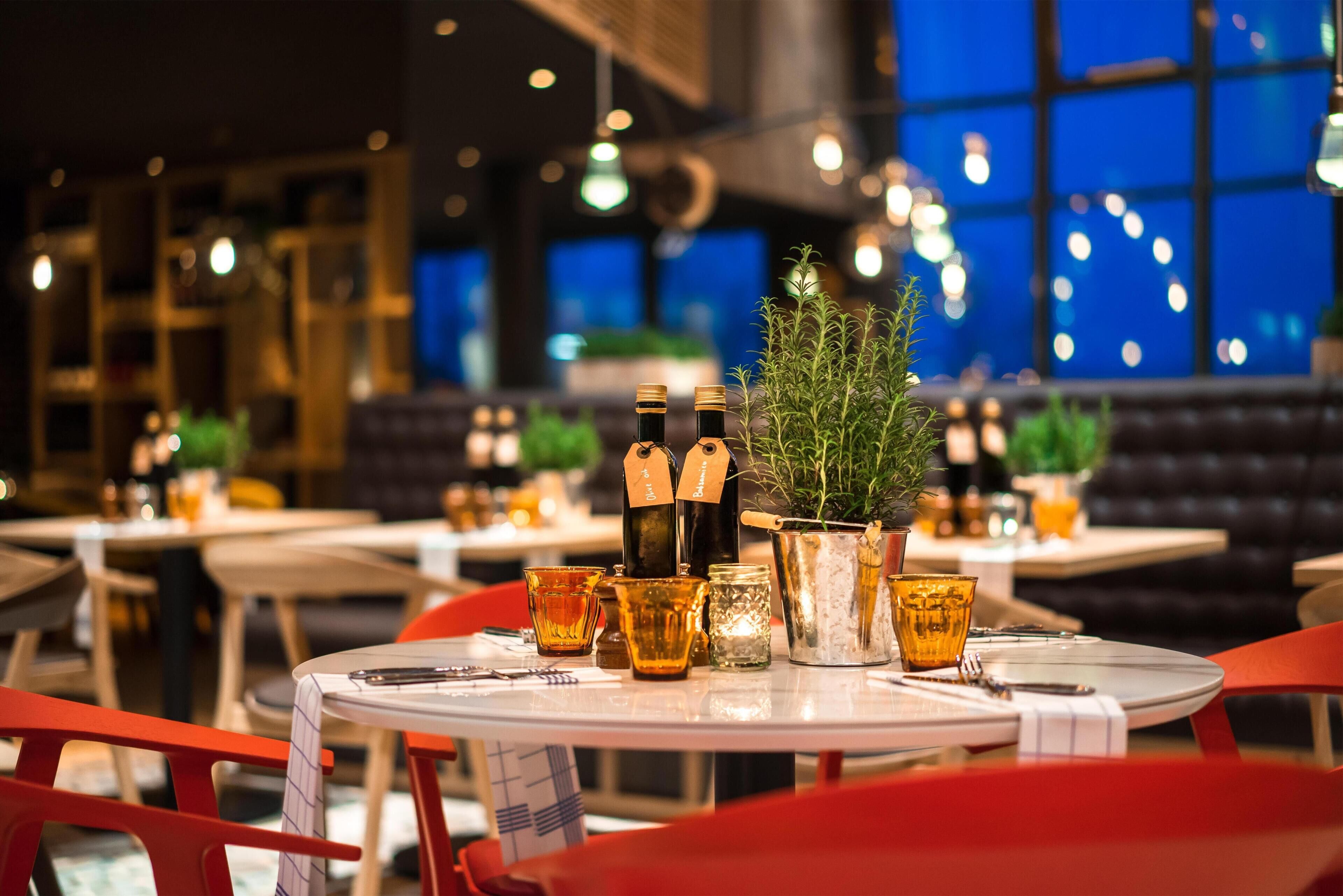 Interior de restaurante con mesas dispuestas para comer, con una planta en maceta y botellas sobre la mesa.