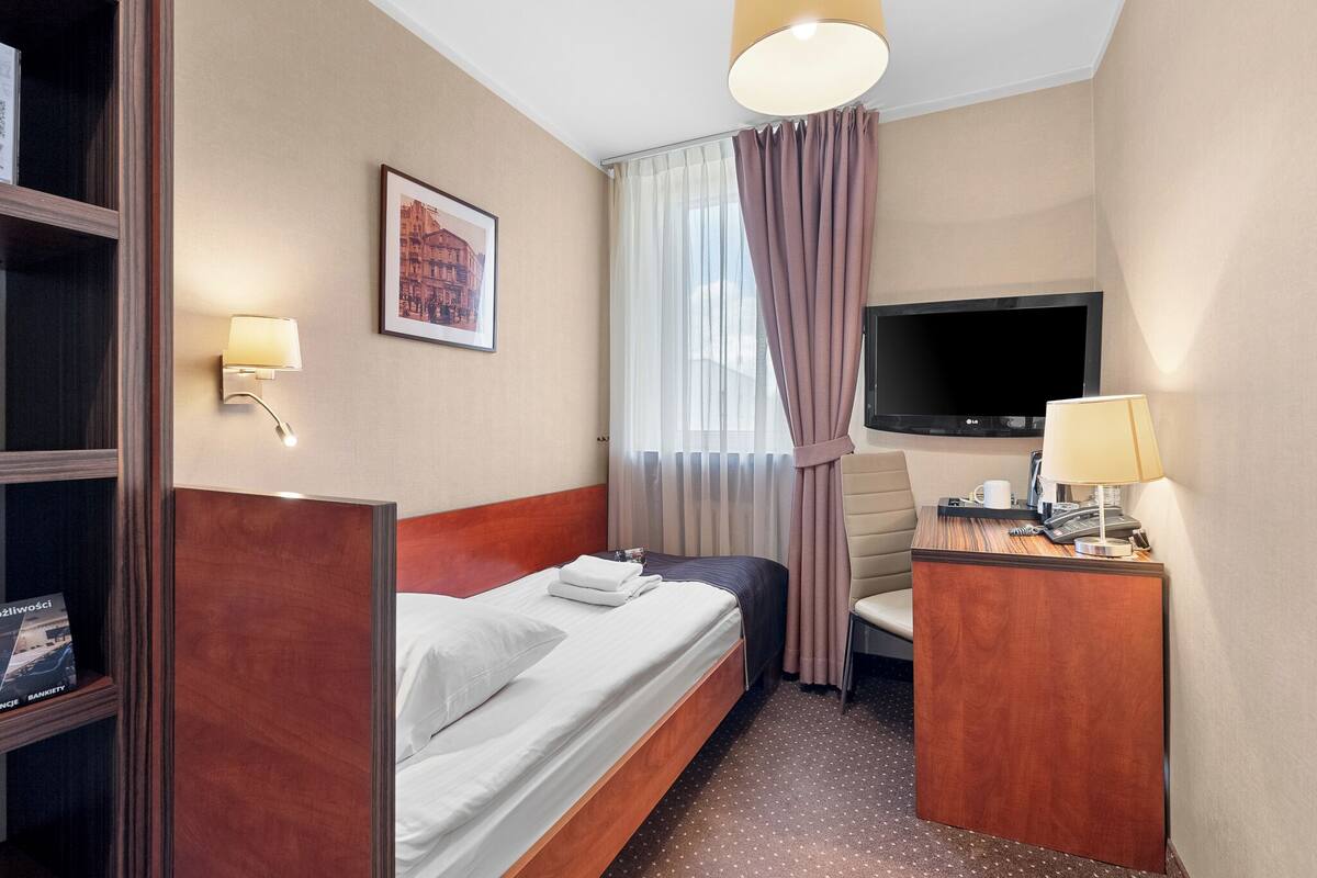 A hotel room with a bed, a desk, a television, a lamp, a chair, a bookshelf, and a framed picture on the wall.