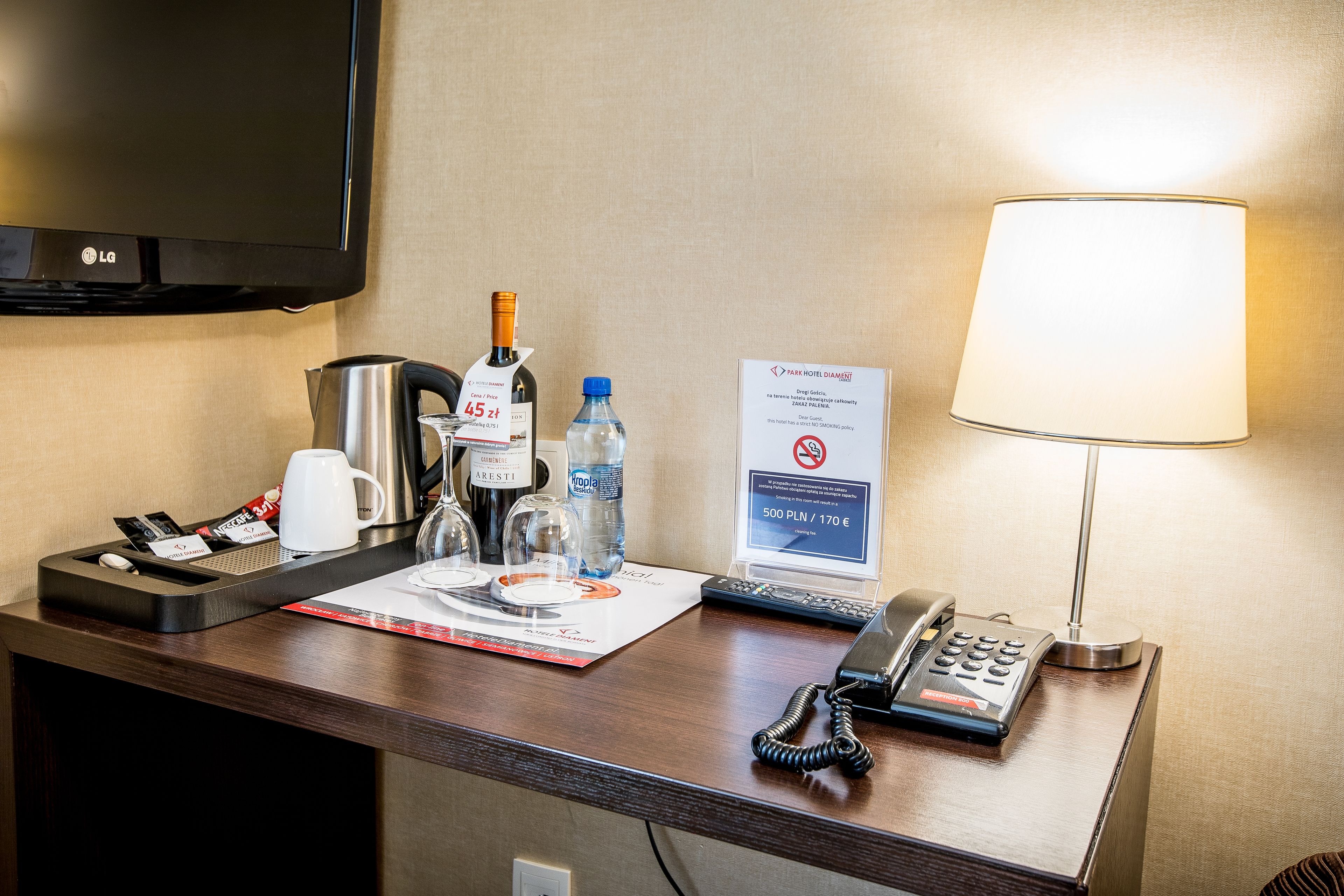 A hotel reception desk with a flat-screen TV, a coffee maker, a bottle of water, a wine bottle, a glass, a telephone, and a sign indicating room rates.