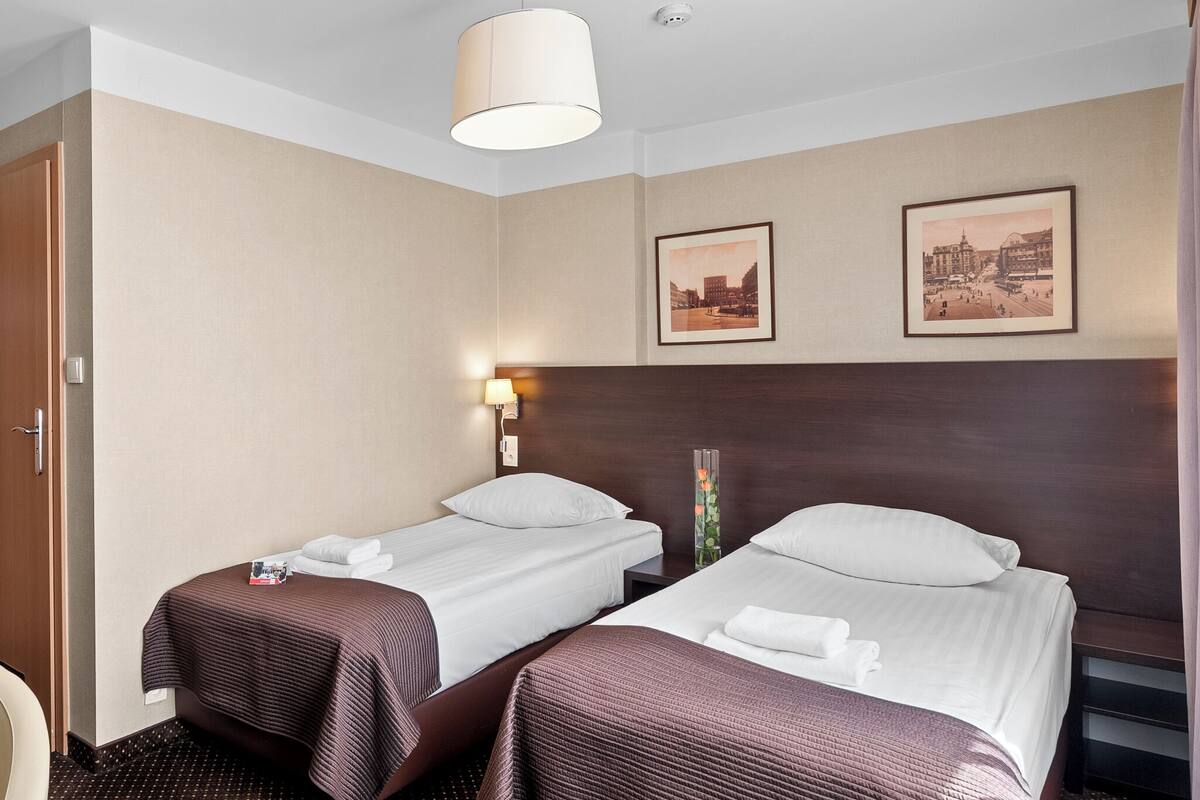 A hotel room with two single beds, a wooden headboard, a wall-mounted lamp, and two framed pictures.