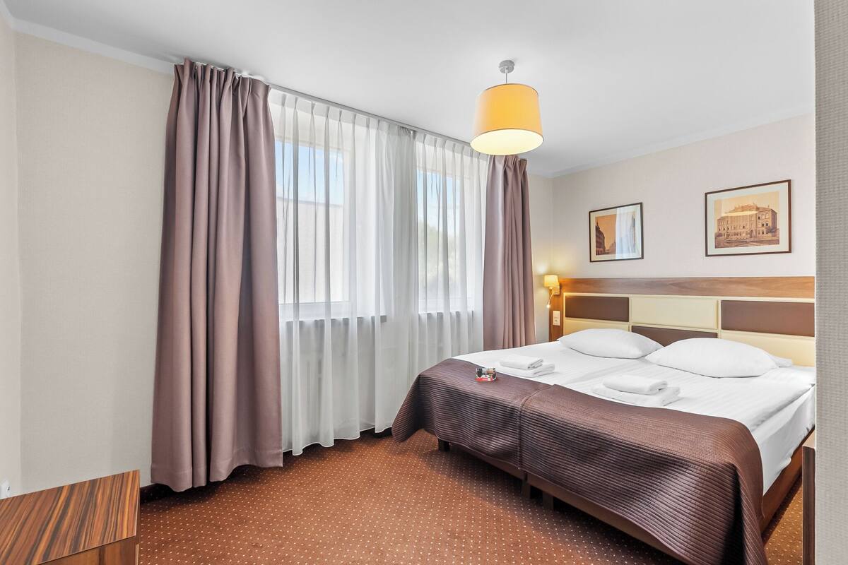A hotel room with a large bed, a wooden nightstand, a lamp, and two framed pictures on the wall.