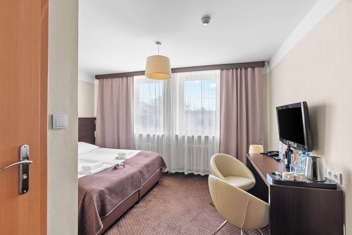 A hotel room with a bed, a desk with a television, a chair, and a window with curtains.