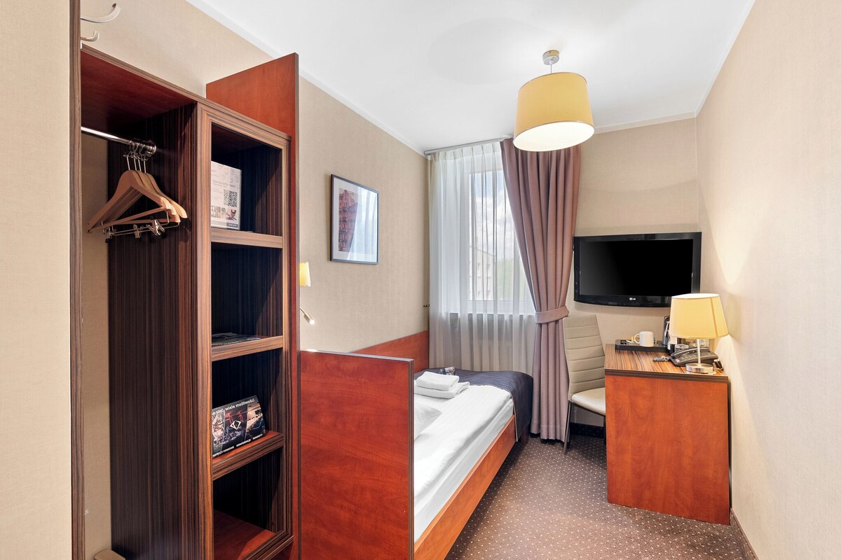 A hotel room with a bed, a desk with a lamp, a TV, a wardrobe, and a window with curtains.