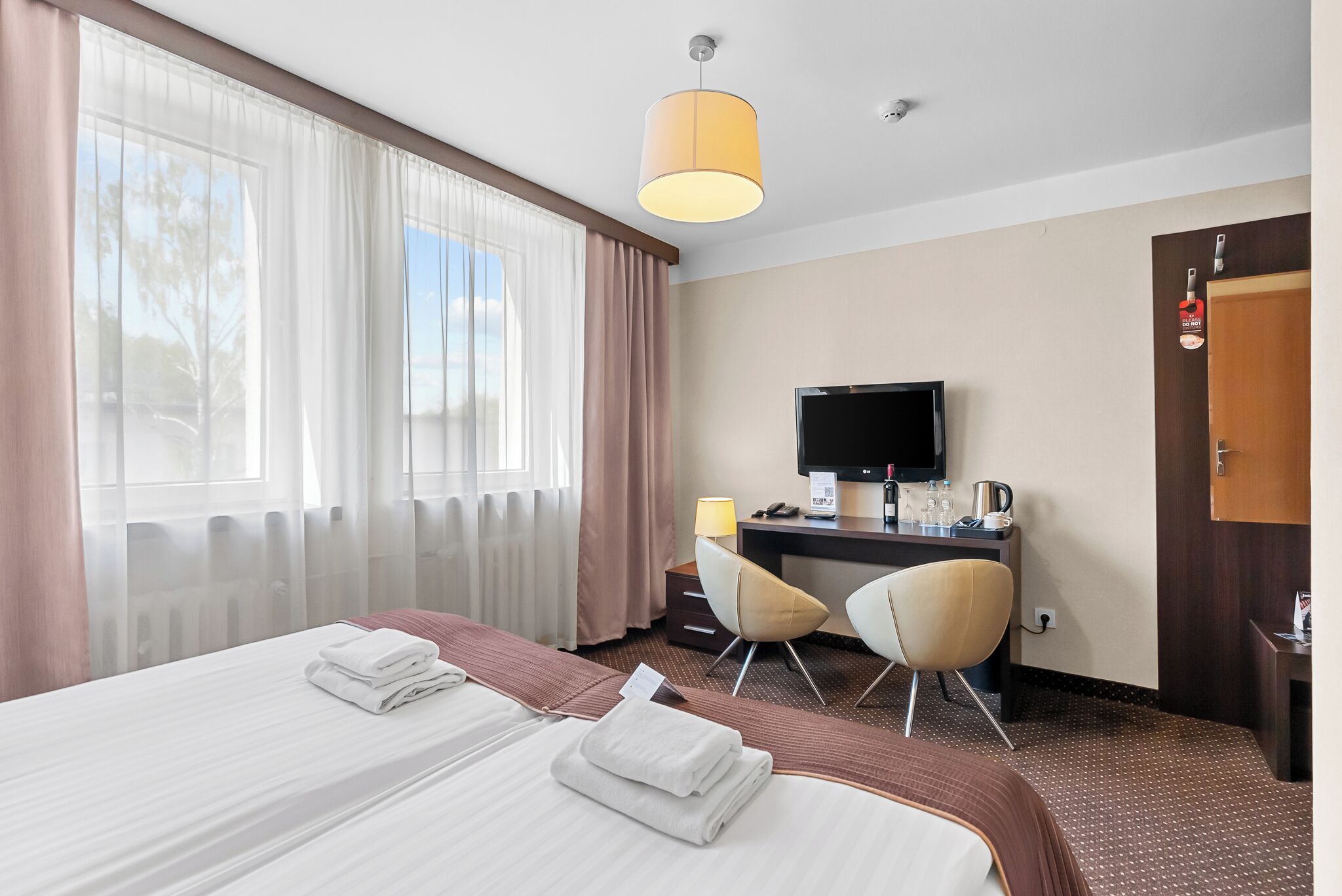 A hotel room with two beds, a desk with a TV, a chair, and a window with curtains.