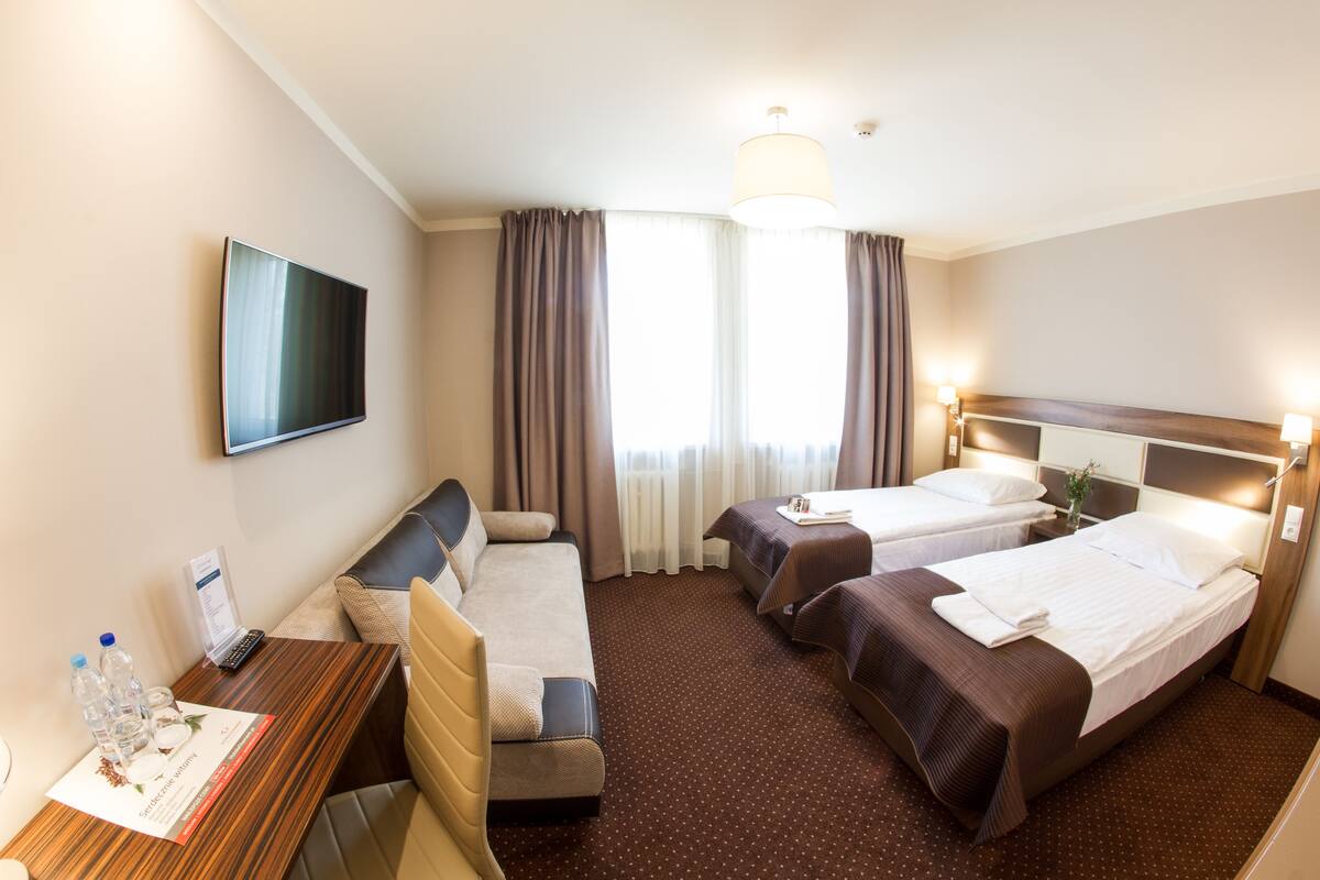 A hotel room with a sofa, two single beds, a desk with a laptop, and a TV on the wall.