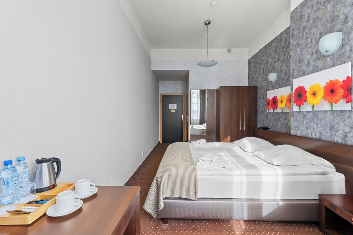 Une chambre d’hôtel avec un lit, une table de chevet en bois, un plateau avec des tasses et une bouilloire, et un tableau représentant des fleurs accroché au mur.