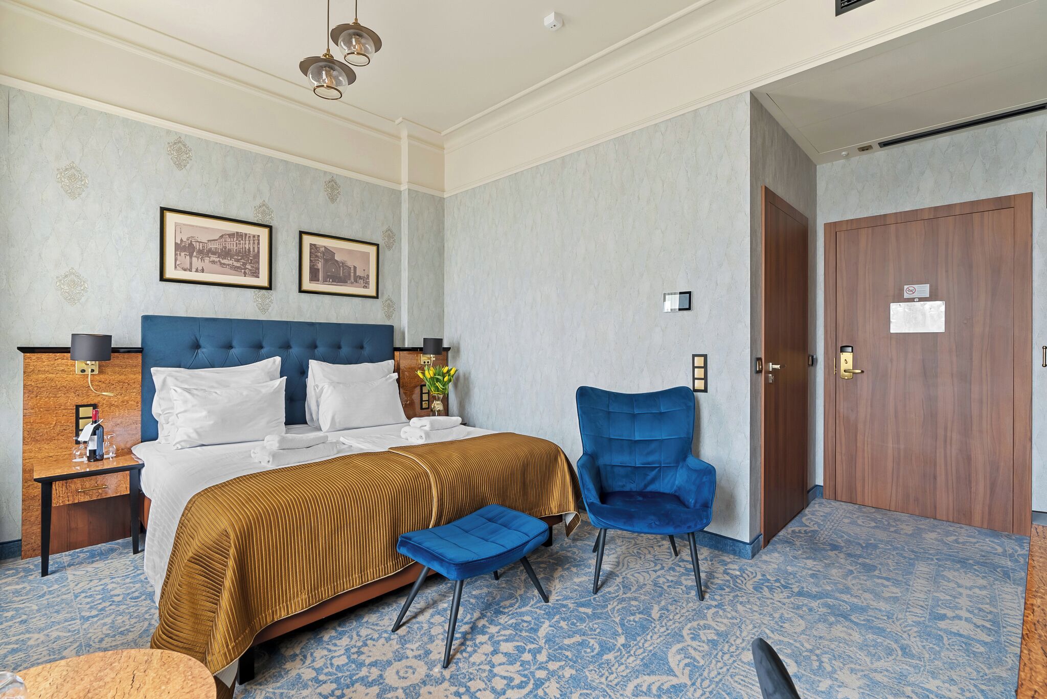 A hotel room with a large bed, a blue armchair, a wooden nightstand, and a door with a sign.