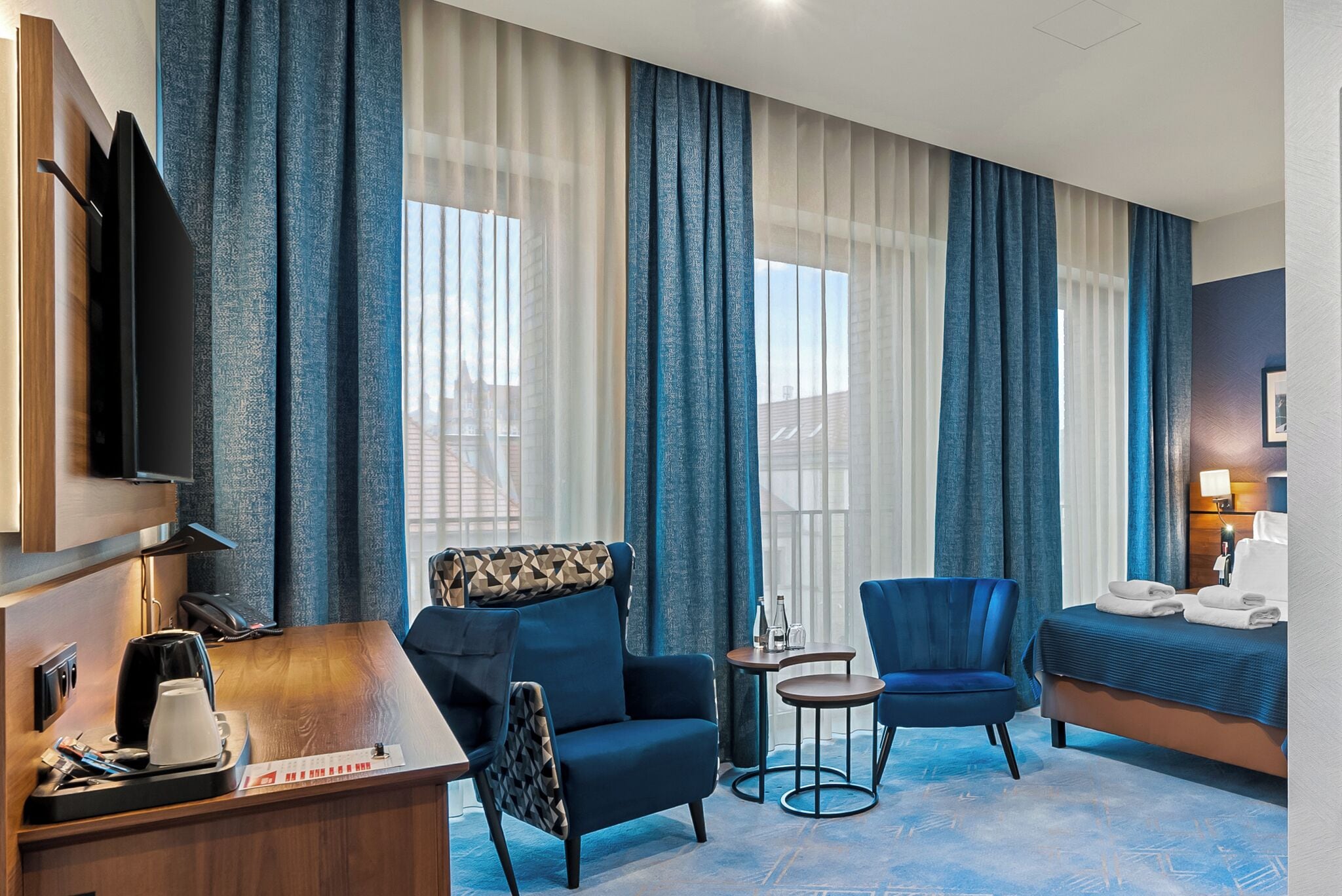 A hotel room with a bed, two armchairs, a small table, a desk with a coffee maker, a TV, and large windows with curtains.