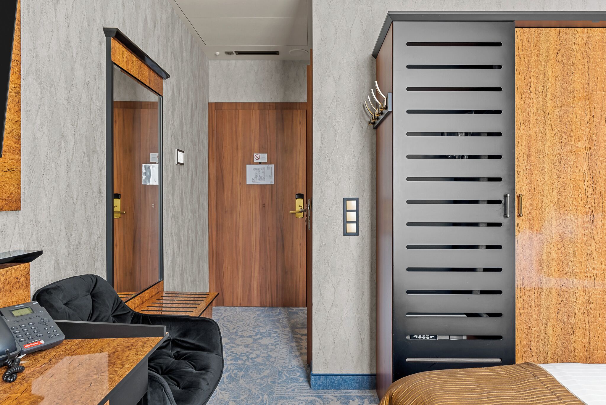 Una camera d'albergo con una porta in legno, un telefono nero, una sedia nera, un letto e un tappeto blu con motivo.
