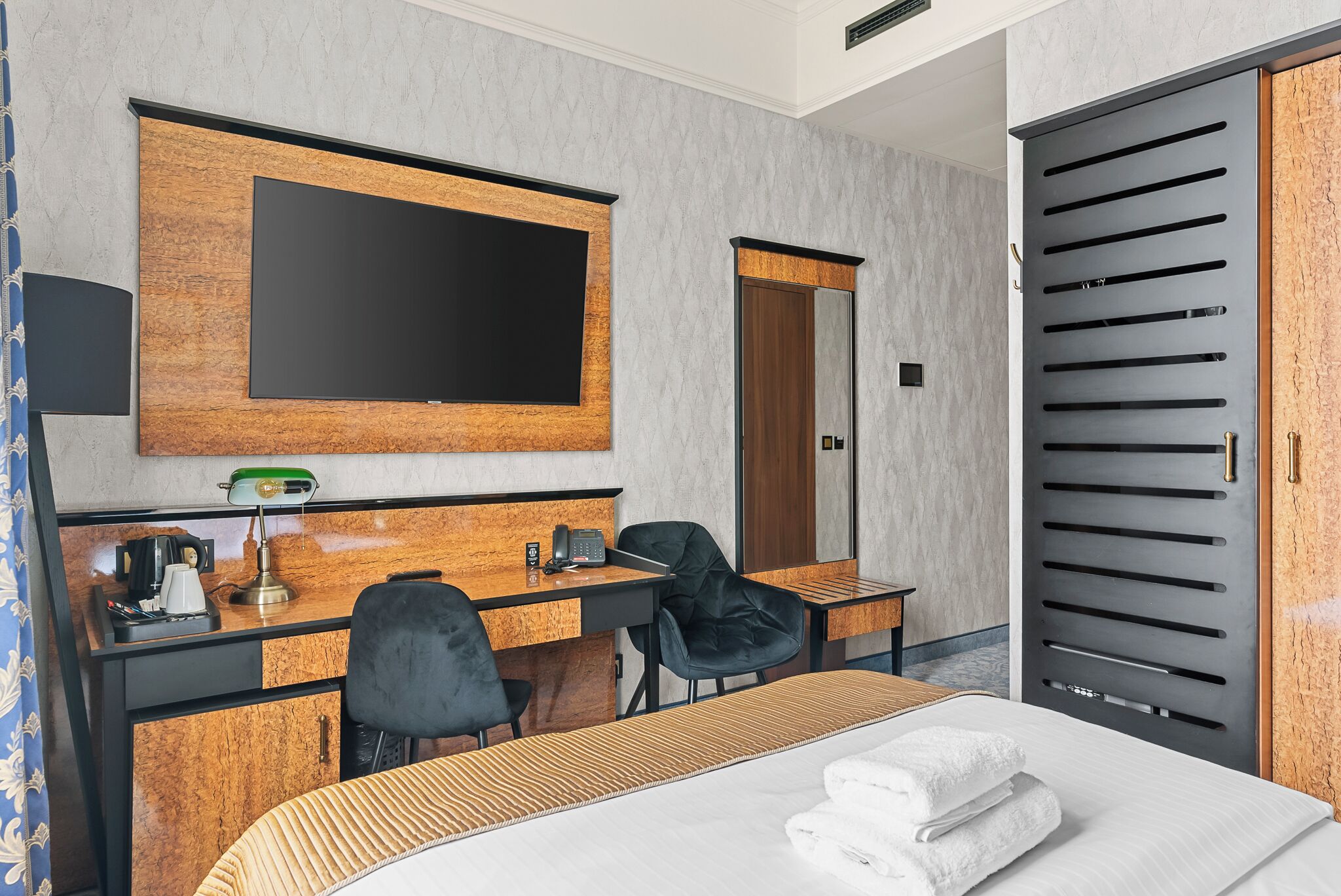 A hotel room with a bed, a desk with a chair, a television, a mirror, and a wardrobe.