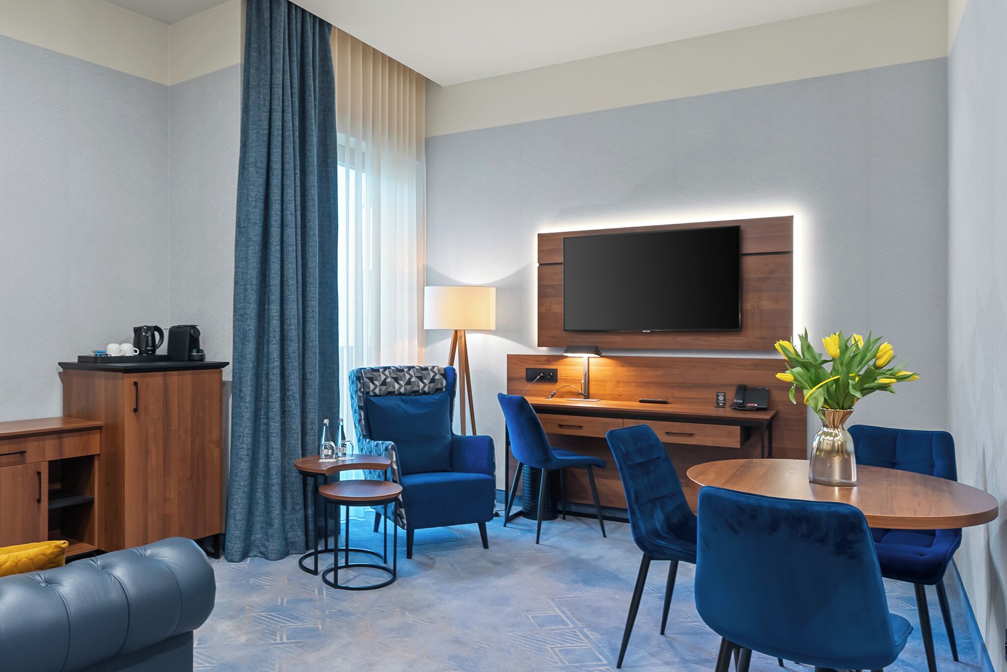 A hotel room with a flat-screen TV, a wooden desk, a blue sofa, and a dining table with blue chairs.