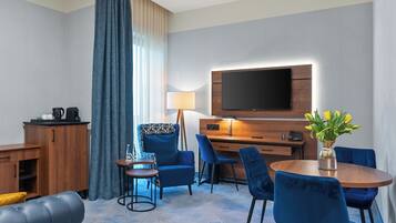 A hotel room with a flat-screen TV, a wooden desk, a blue sofa, and a dining table with blue chairs.