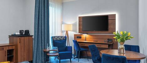 Habitación de hotel con televisor de pantalla plana, escritorio de madera, sofá azul y mesa de comedor con sillas azules.