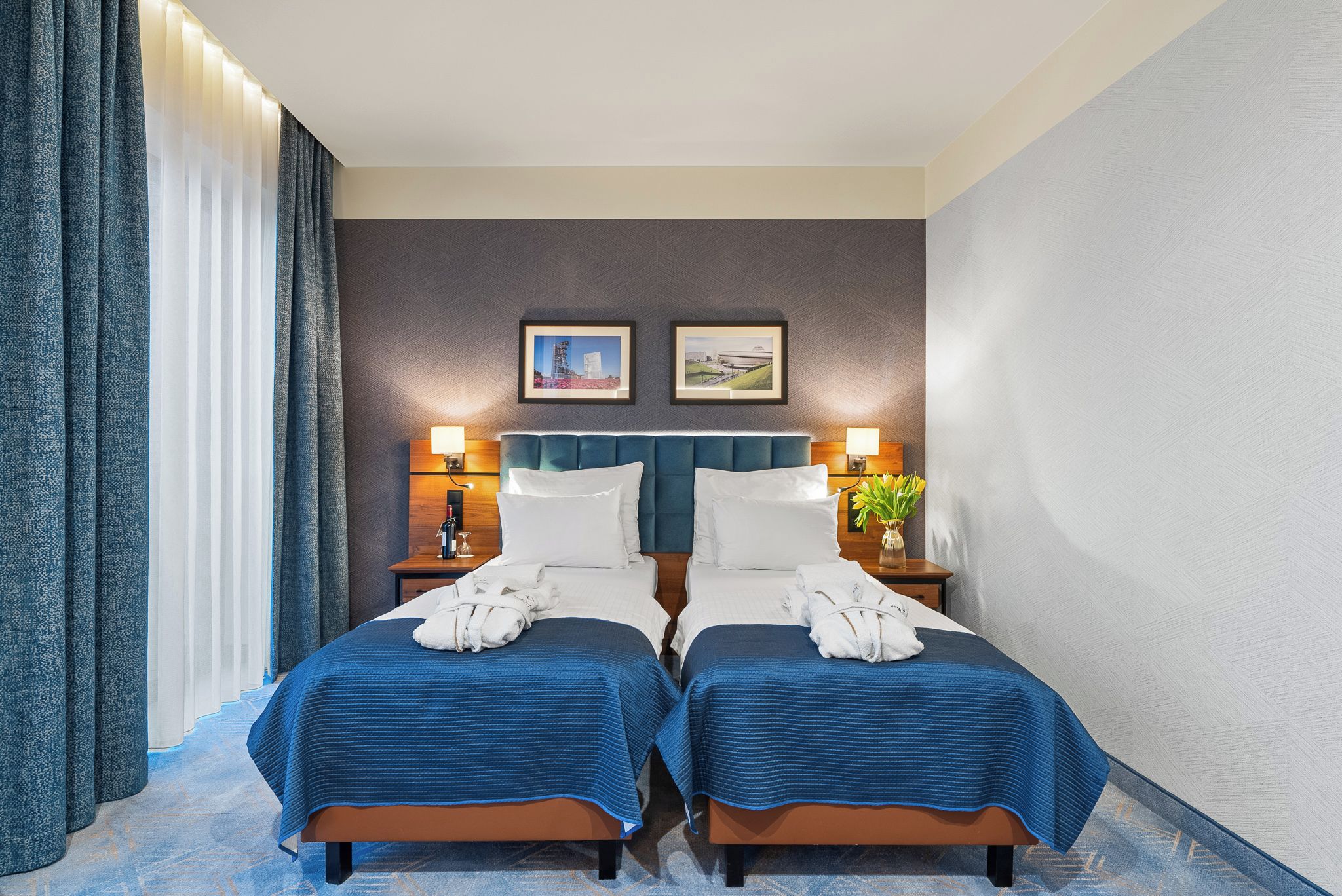 A hotel room with two beds, a large window with curtains, a wall-mounted lamp, and two framed pictures.